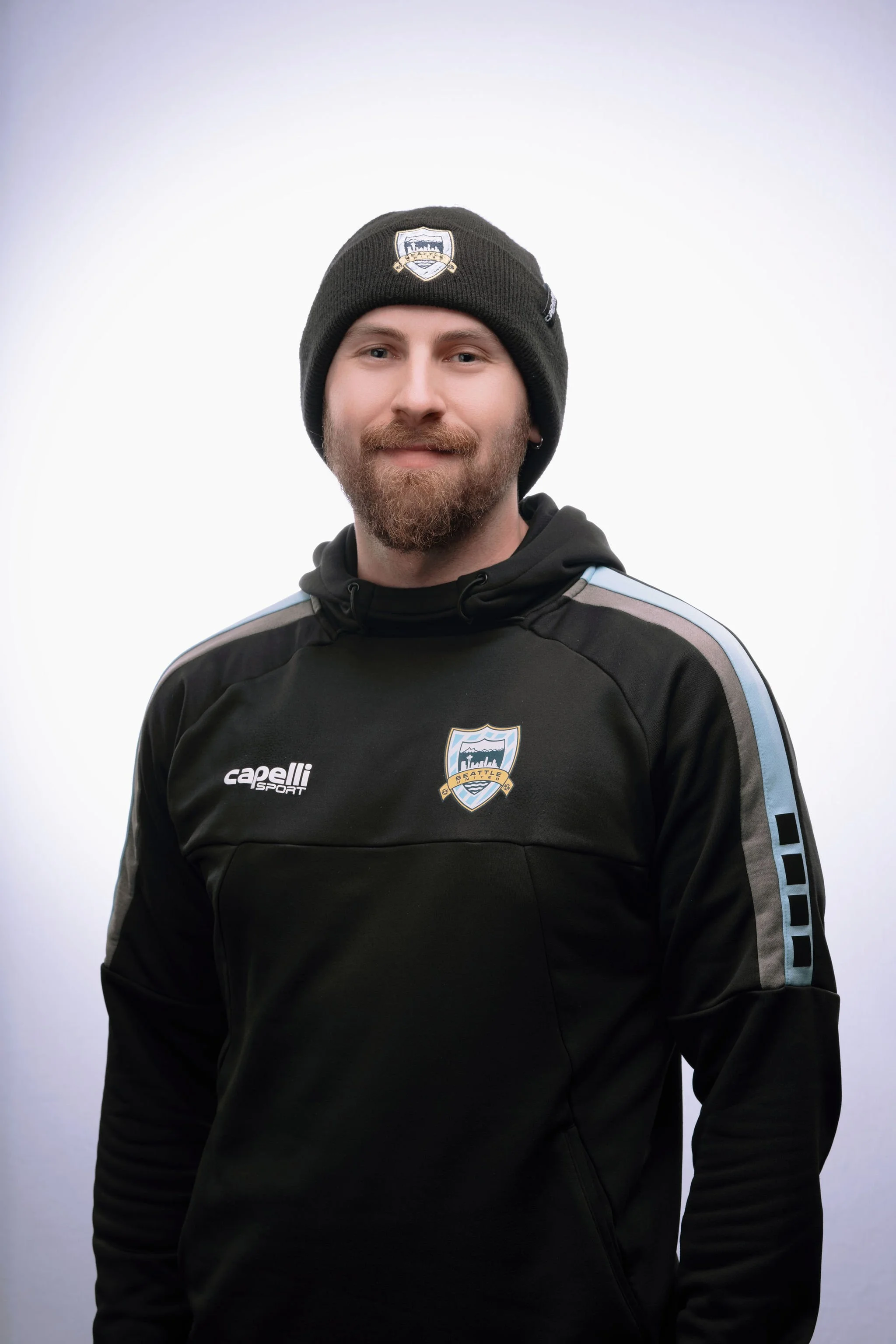 A man wearing a black hoodie and a black beanie with a Seattle soccer team logo, standing against a plain white background.