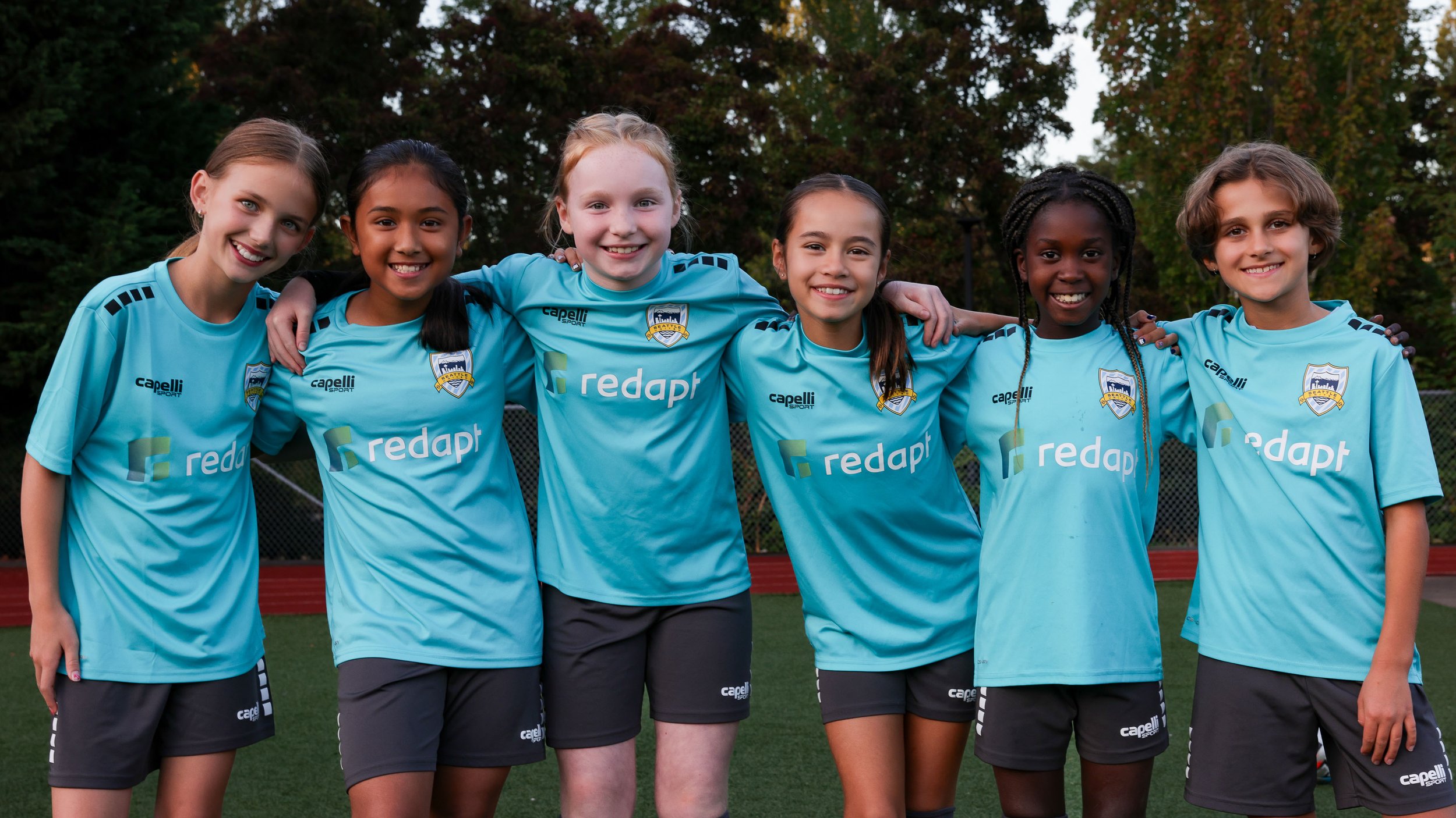Group of six young girls in light blue soccer uniforms, standing outdoors with their arms around each other's shoulders, smiling at the camera.