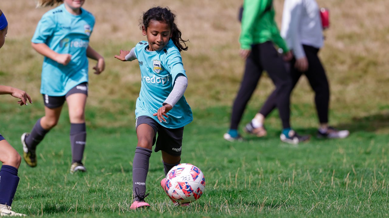 Young girl in a blue soccer uniform kicking a soccer ball on a grassy field with other children and adults in the background.