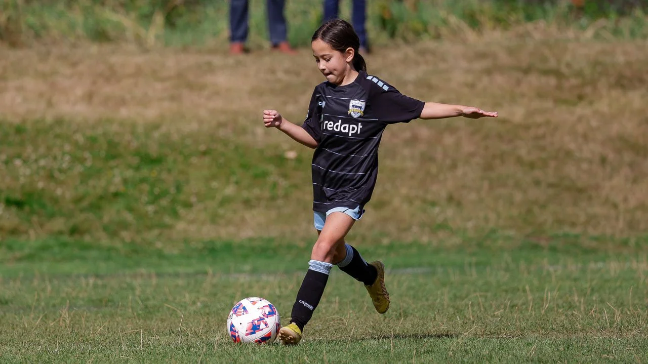 A young girl in a soccer uniform kicks a ball on a grassy field.