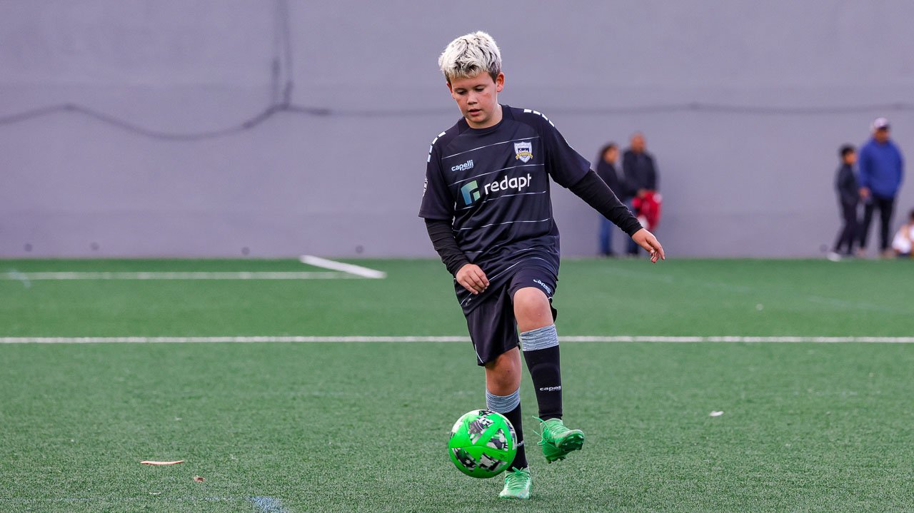 Young boy playing soccer on a field with a green ball, wearing a black sports uniform.