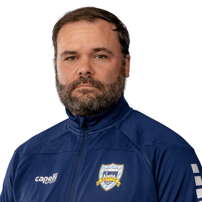 A man with short brown hair and a beard wearing a navy blue sports jacket with the Seattle Sounders FC logo.