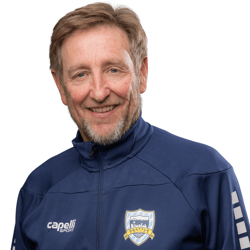 A smiling man with light brown hair and a beard, wearing a blue sports jacket with a Seattle sports team logo and the brand name Capelli Sport, looking directly at the camera.