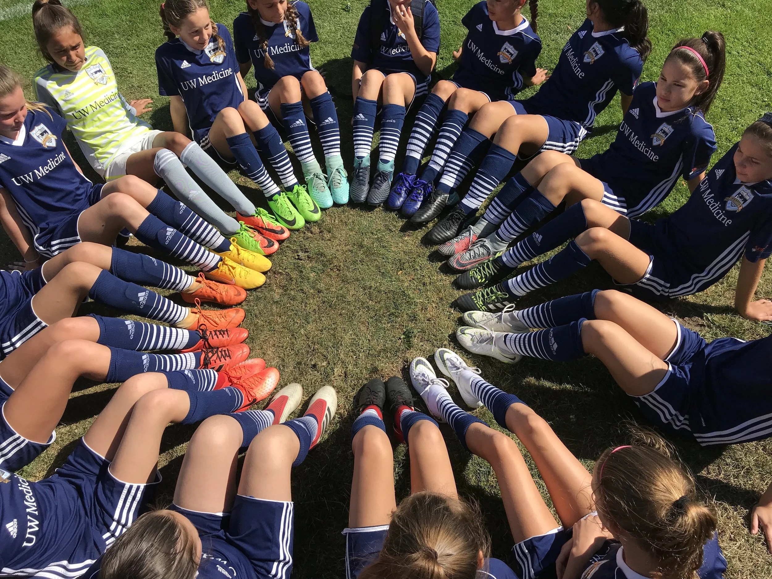 A group of young female soccer players from UW Medicine sitting in a circle on the grass, wearing blue uniforms and colorful cleats, with their legs extended inward.