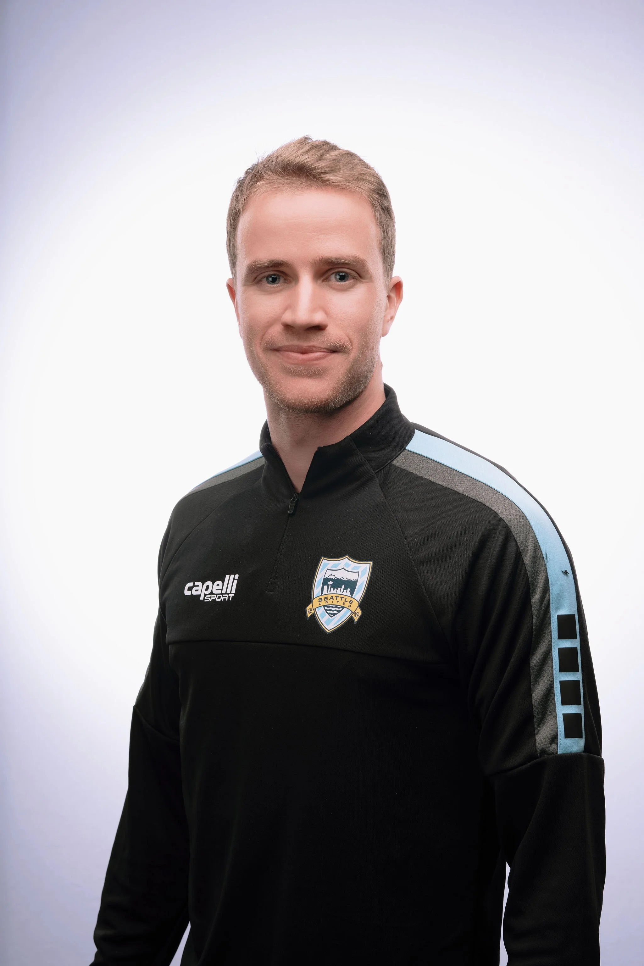 A man wearing a black Seahawks athletic jacket with the team logo on the chest, standing against a light gradient background.