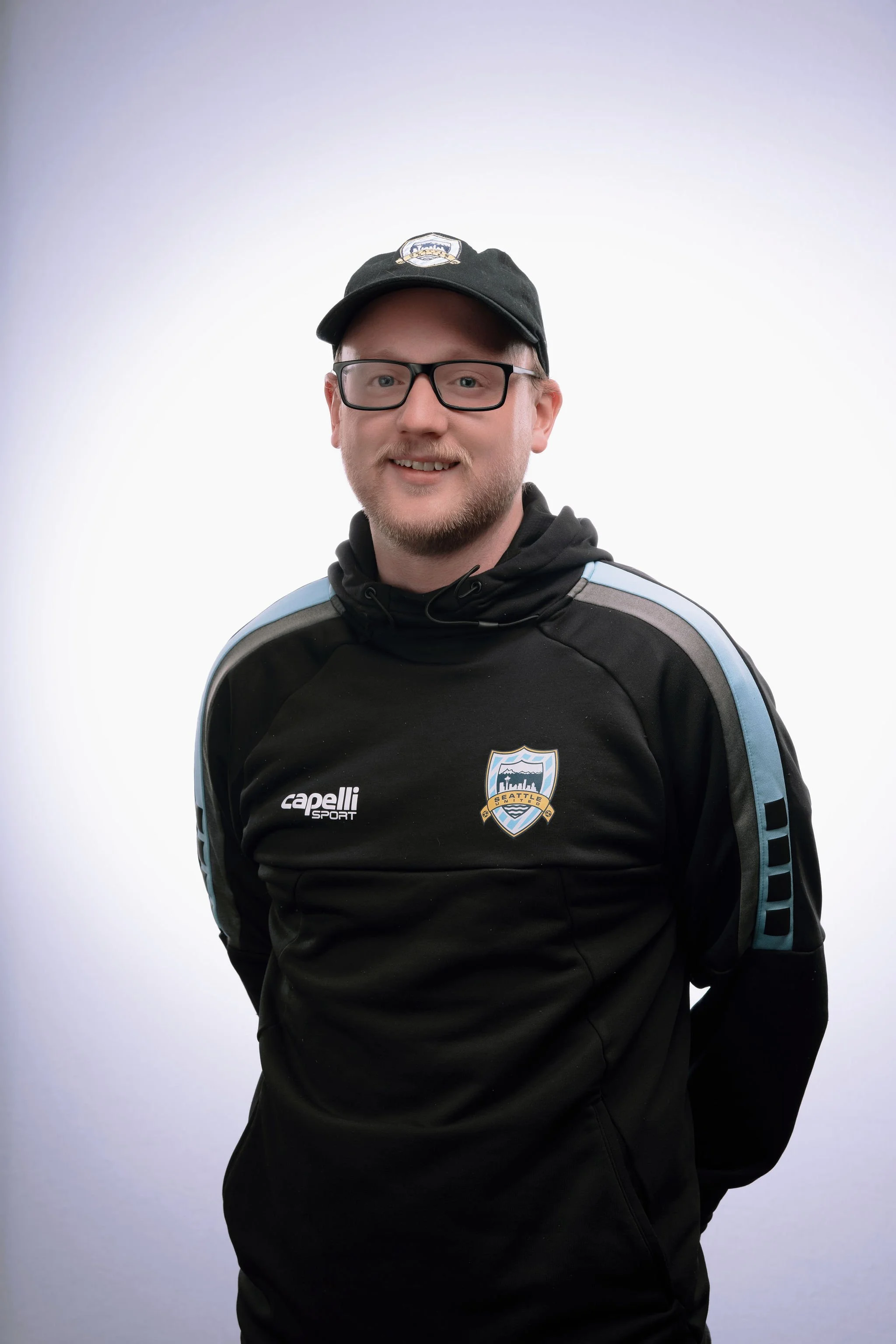 A young man with glasses and a beard, smiling, wearing a black Seattle soccer jacket with a cap that has a Seattle logo, standing in front of a plain white background.
