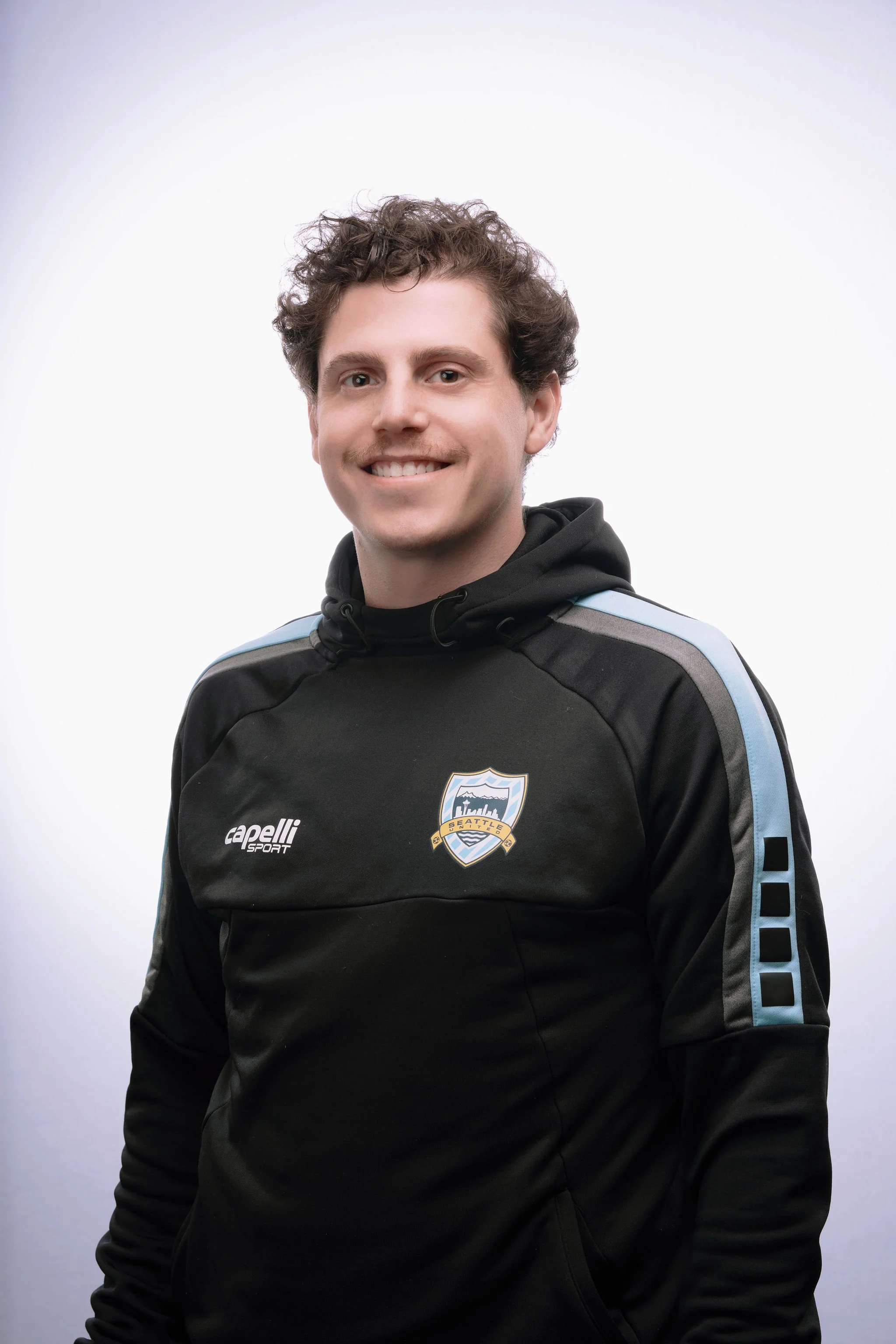 A young man with curly brown hair and a smile, wearing a black sports hoodie with a Seattle sports team logo on the chest and light blue and gray accents on the sleeves, standing against a plain white background.