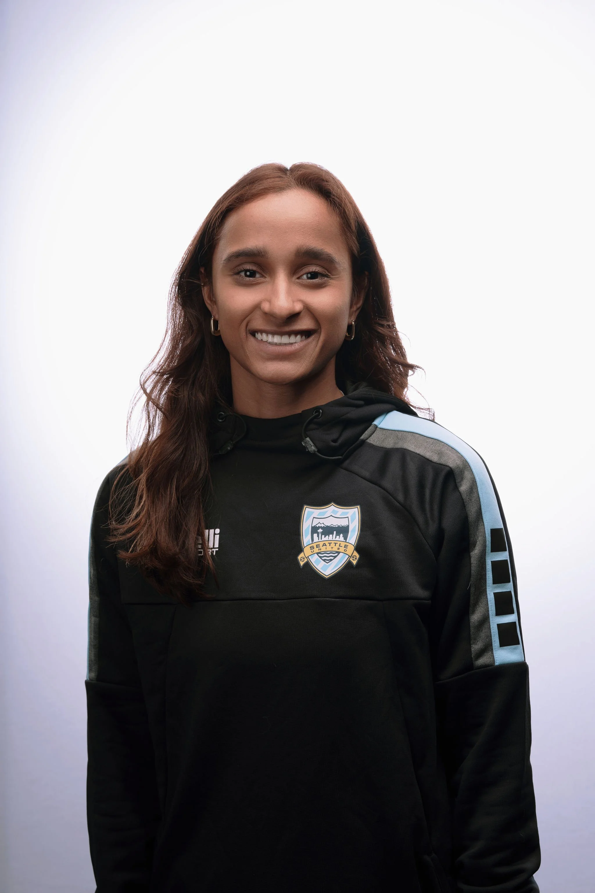 A woman smiling at the camera wearing a black jacket with a Seattle soccer team logo on it, with a white background.