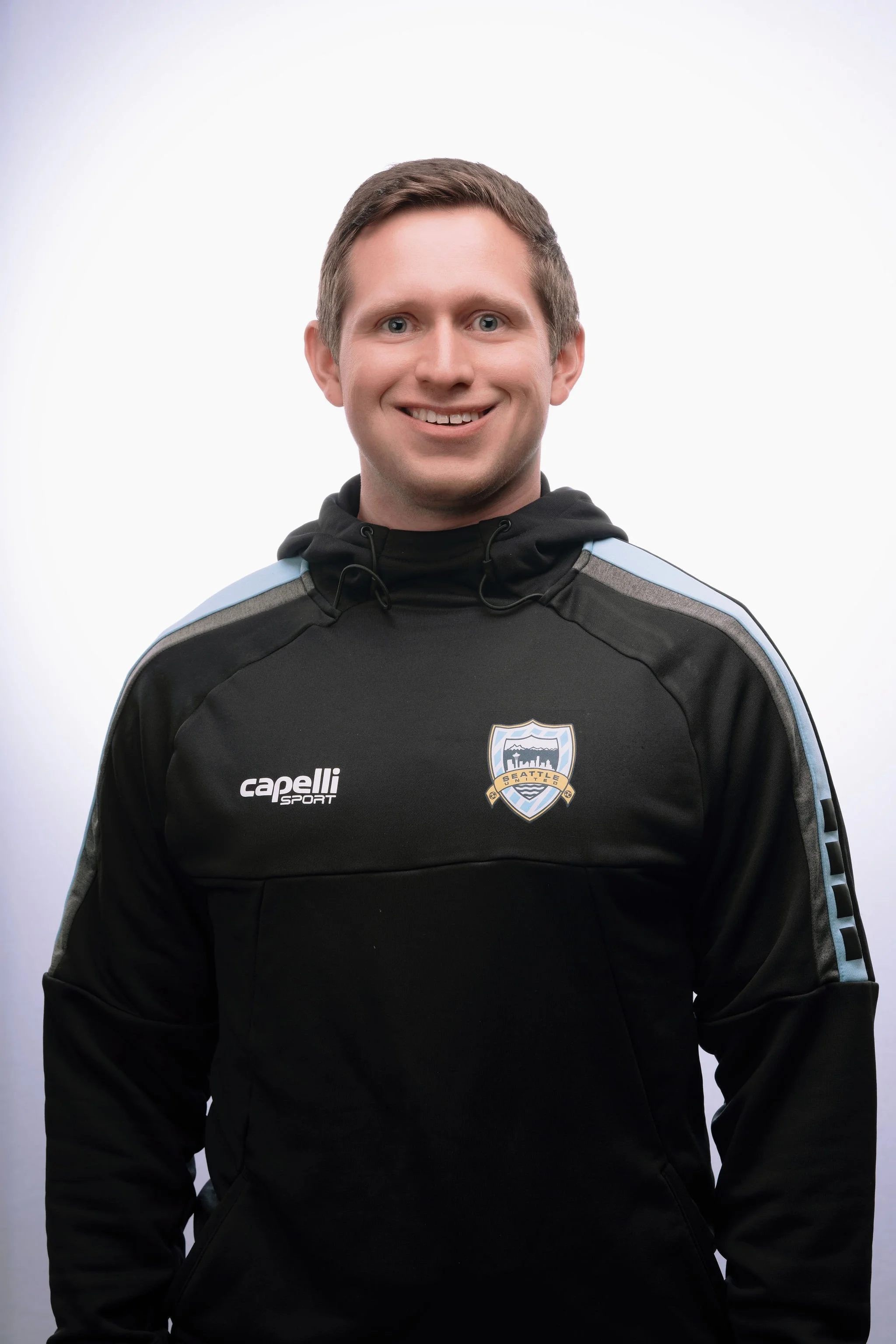 Young man smiling, wearing a black Seattle sports jacket with a Capelli Sport logo, standing against a plain white background.