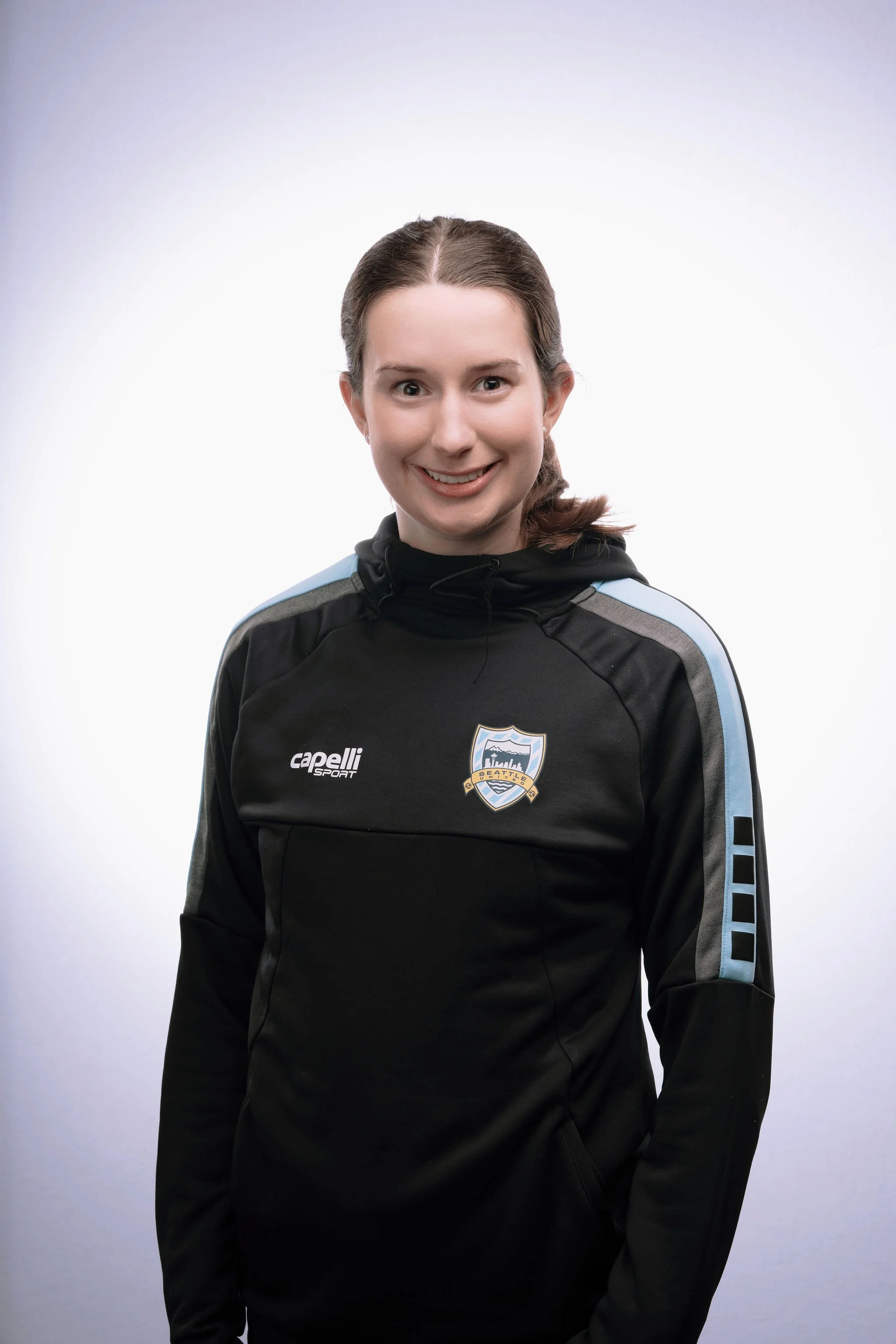 A young woman with brown hair in a ponytail, wearing a black sports jacket with Seattle Sounders FC logo, smiling at the camera, against a plain white background.