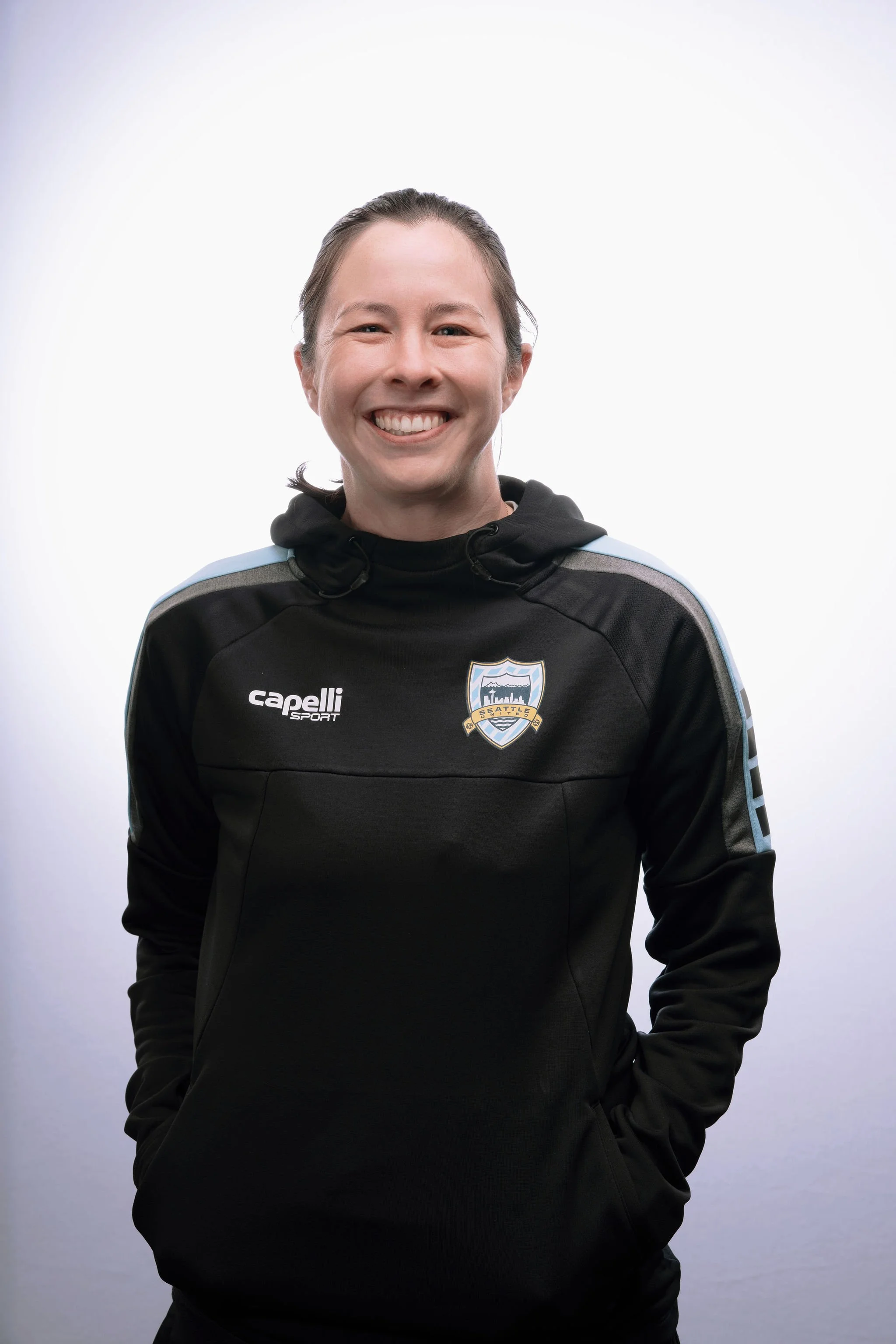 Smiling woman in black sports jacket with team logo, hands in pockets, standing against a white background.