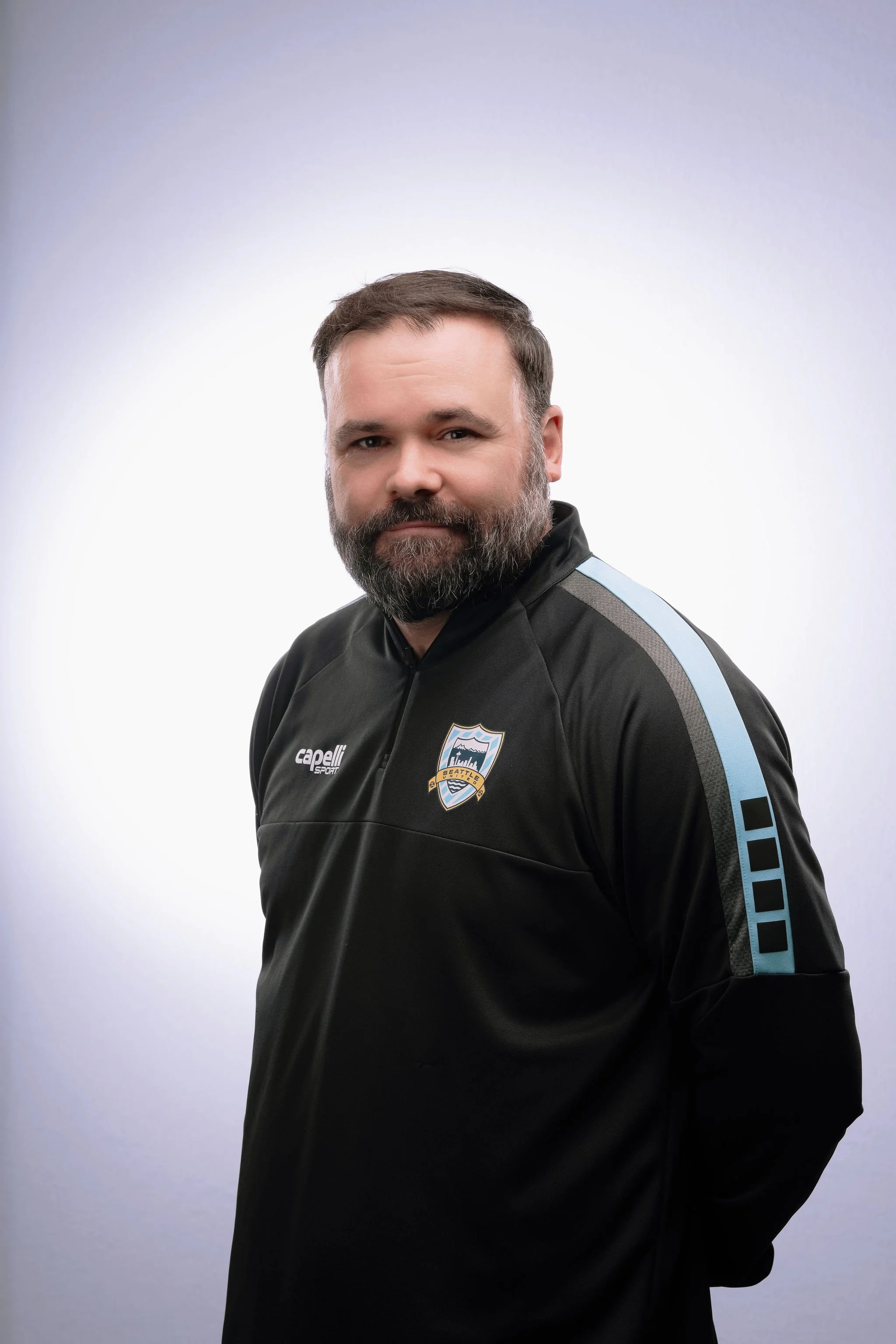 A man with a beard wearing a black athletic jacket with a Seattle Sounders FC badge, posing against a plain white background.