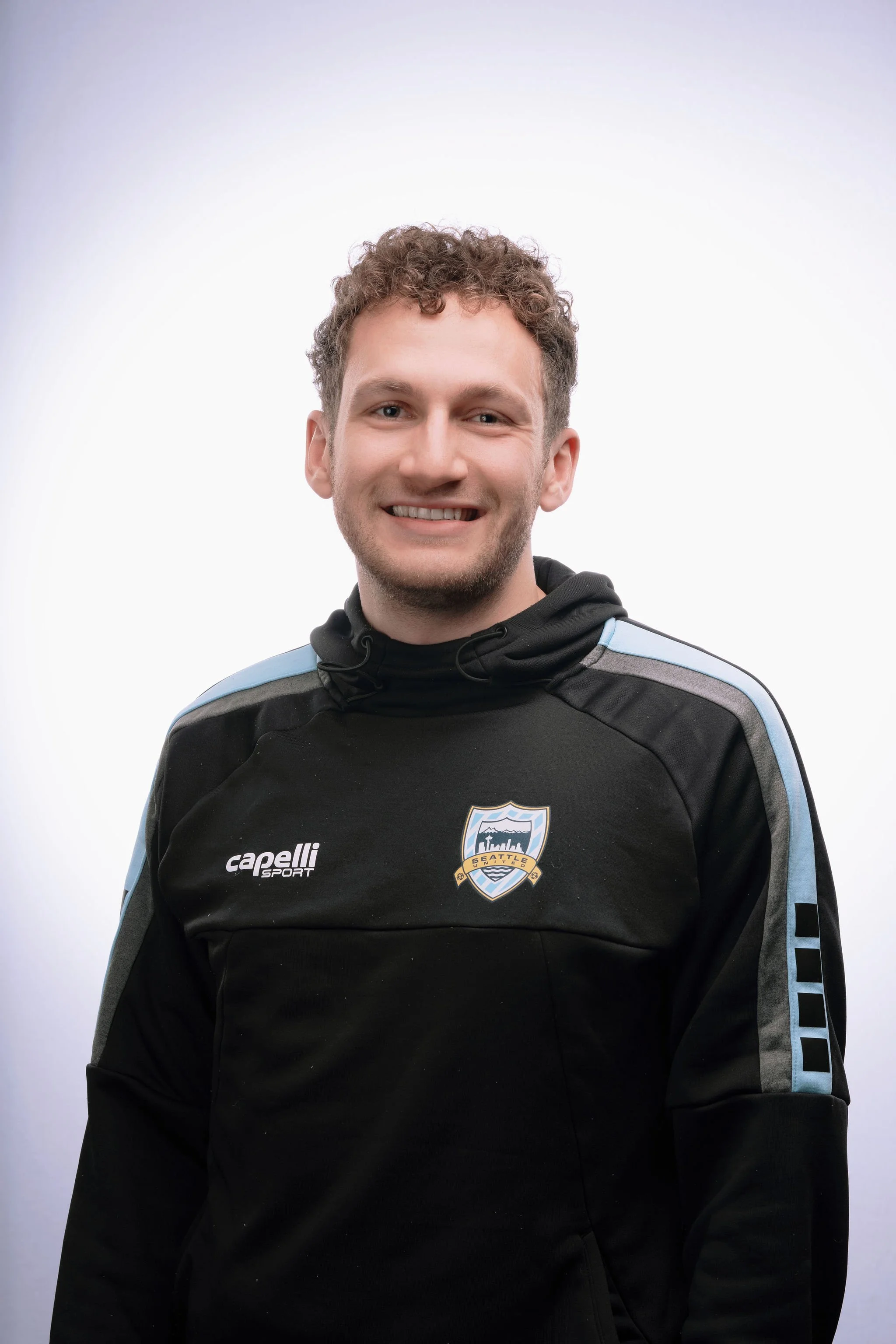 A smiling man wearing a black sports hoodie with Seattle Football Club logo and Capelli Sport branding, standing in front of a plain white background.
