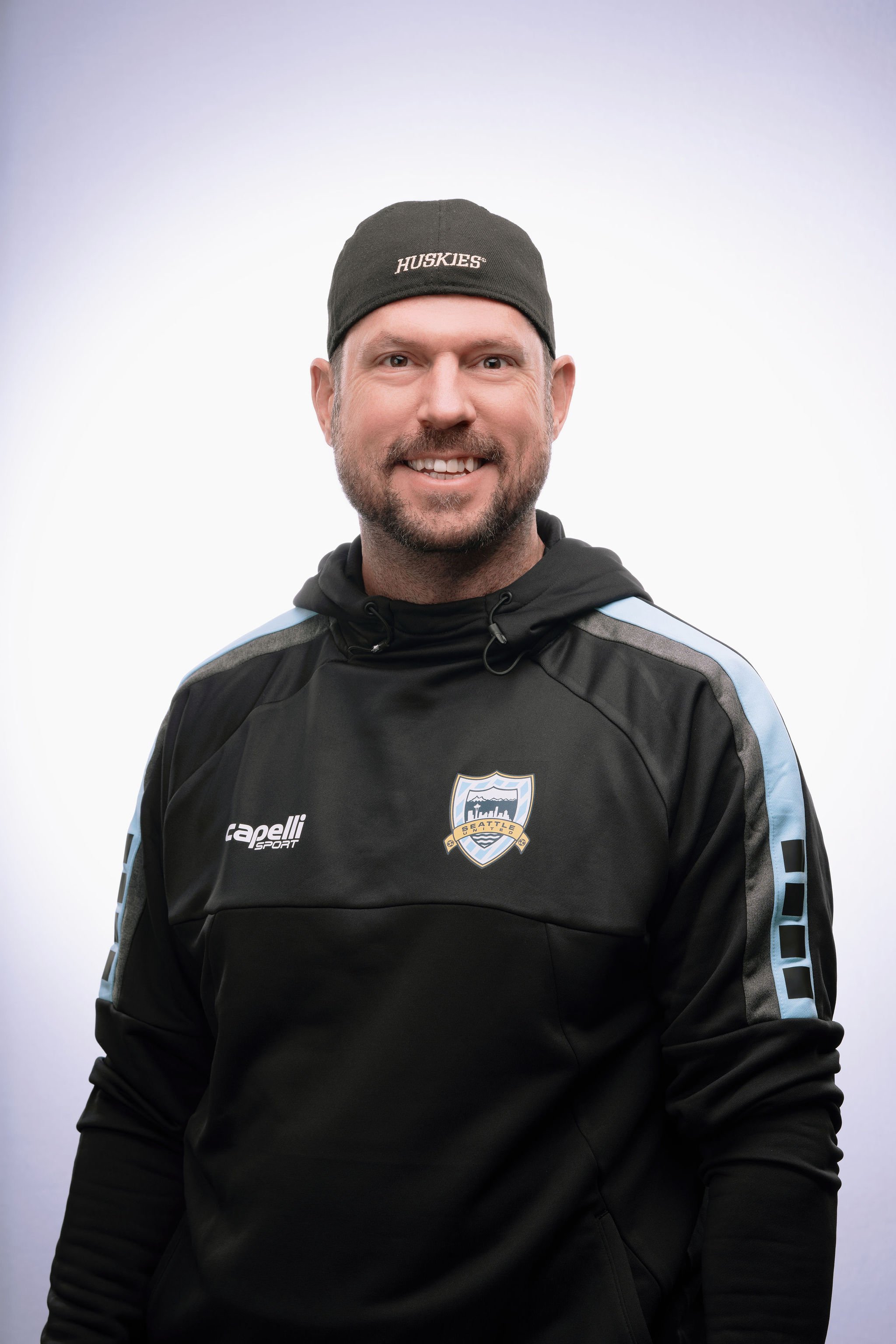 A smiling man wearing a black hoodie with Seattle Reign FC logo and a black beanie with 'Huskies' embroidered on it, standing against a plain light background.