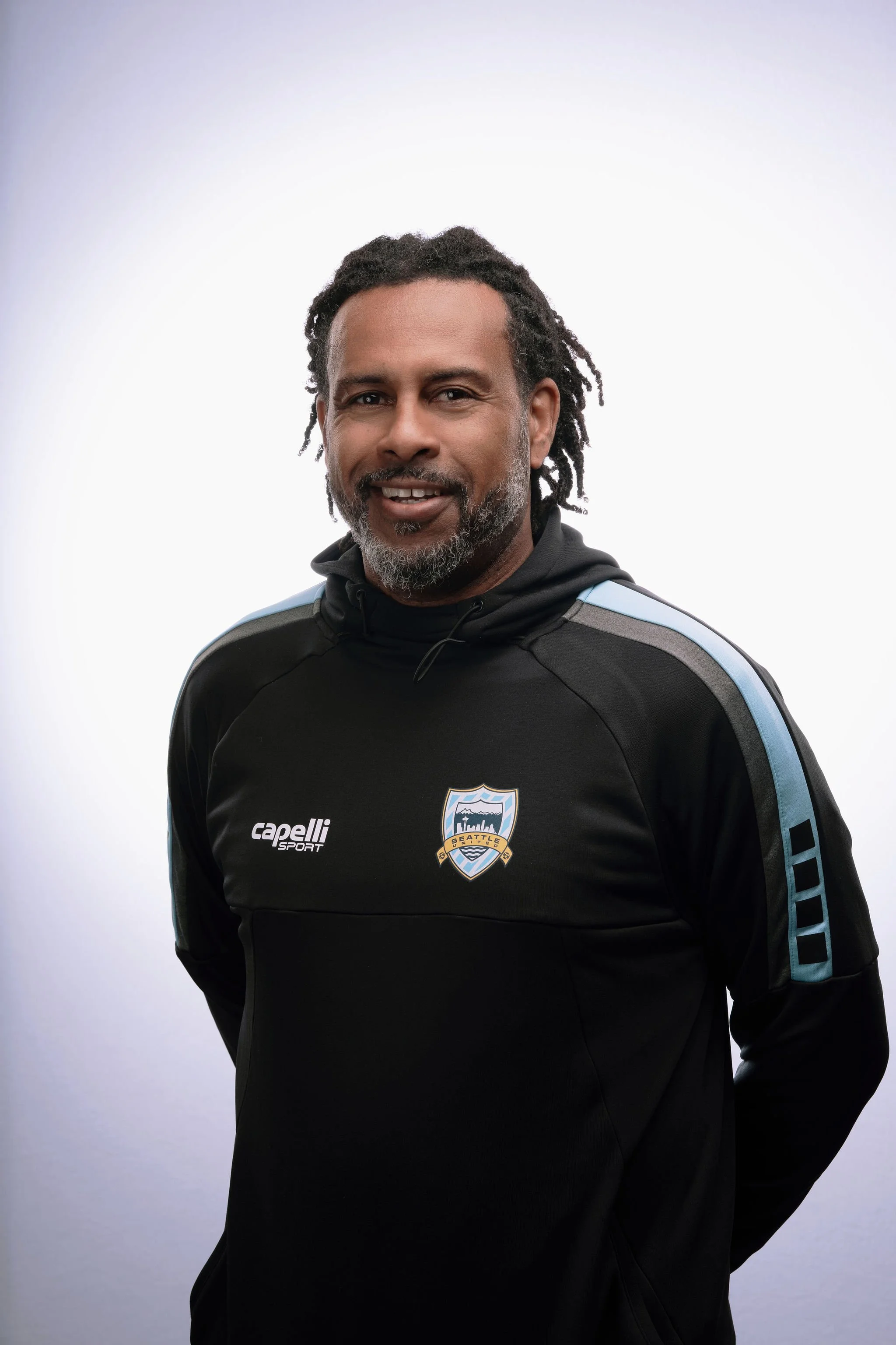 A man with dreadlocks and a beard wearing a black sports jacket with a Seattle team logo and the Capelli Sport brand, standing against a white background.