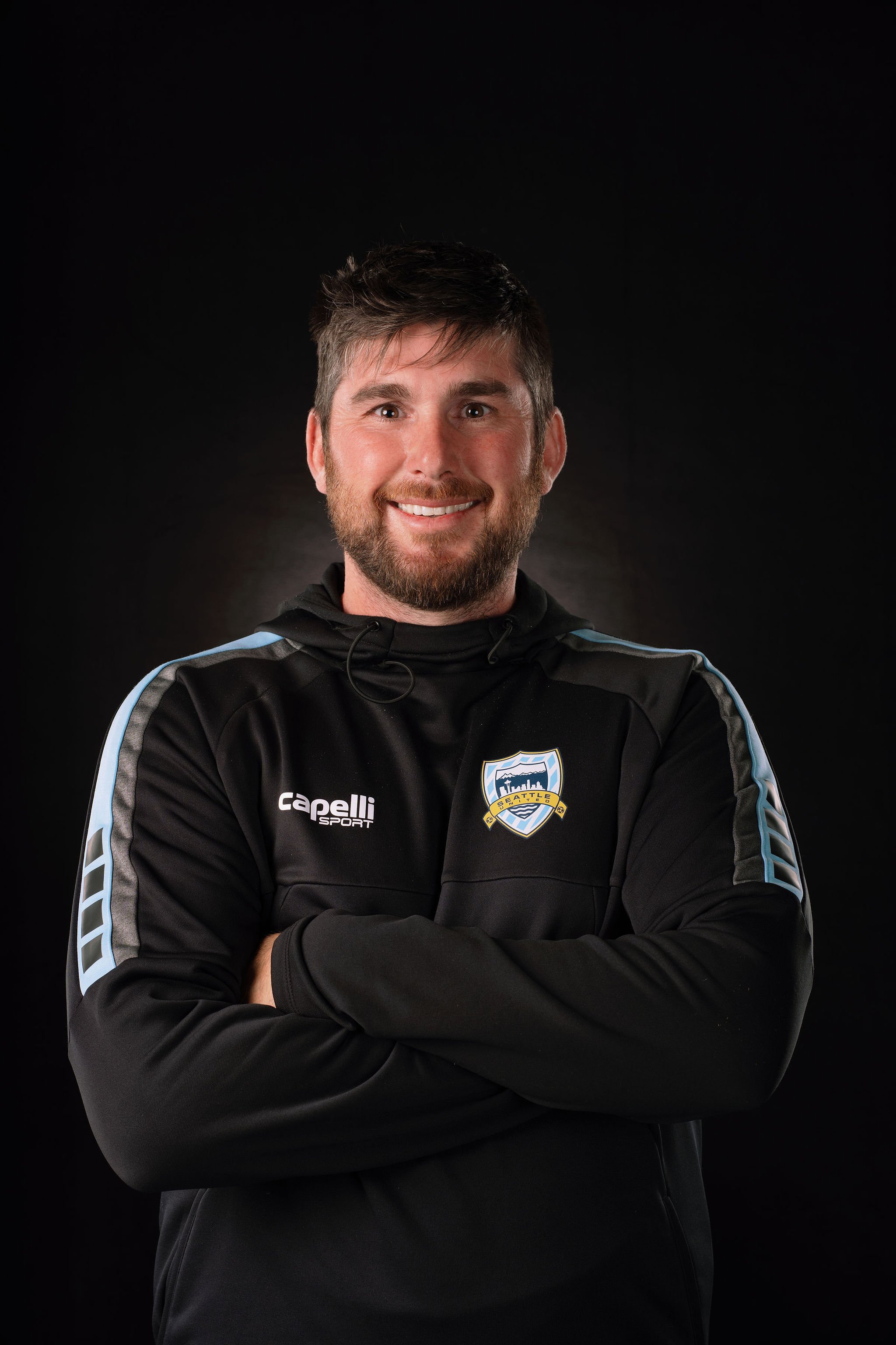 Smiling man with short dark hair and a beard, wearing a black hoodie with a pink and white logo in front, standing against a textured dark wall.