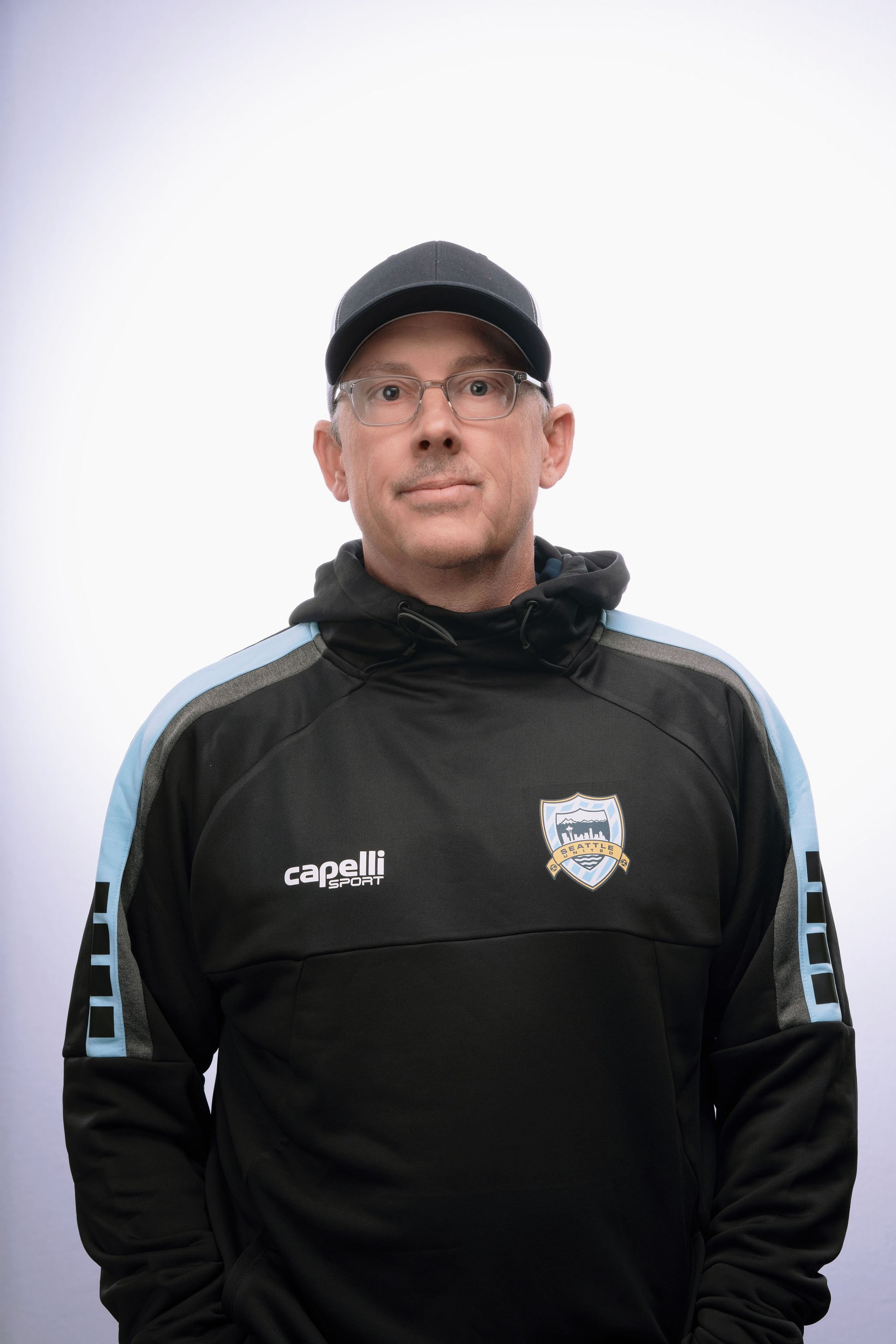 A man wearing a black and light blue sports jacket with a Seattle football team logo, a black cap, and glasses, standing against a plain white background.