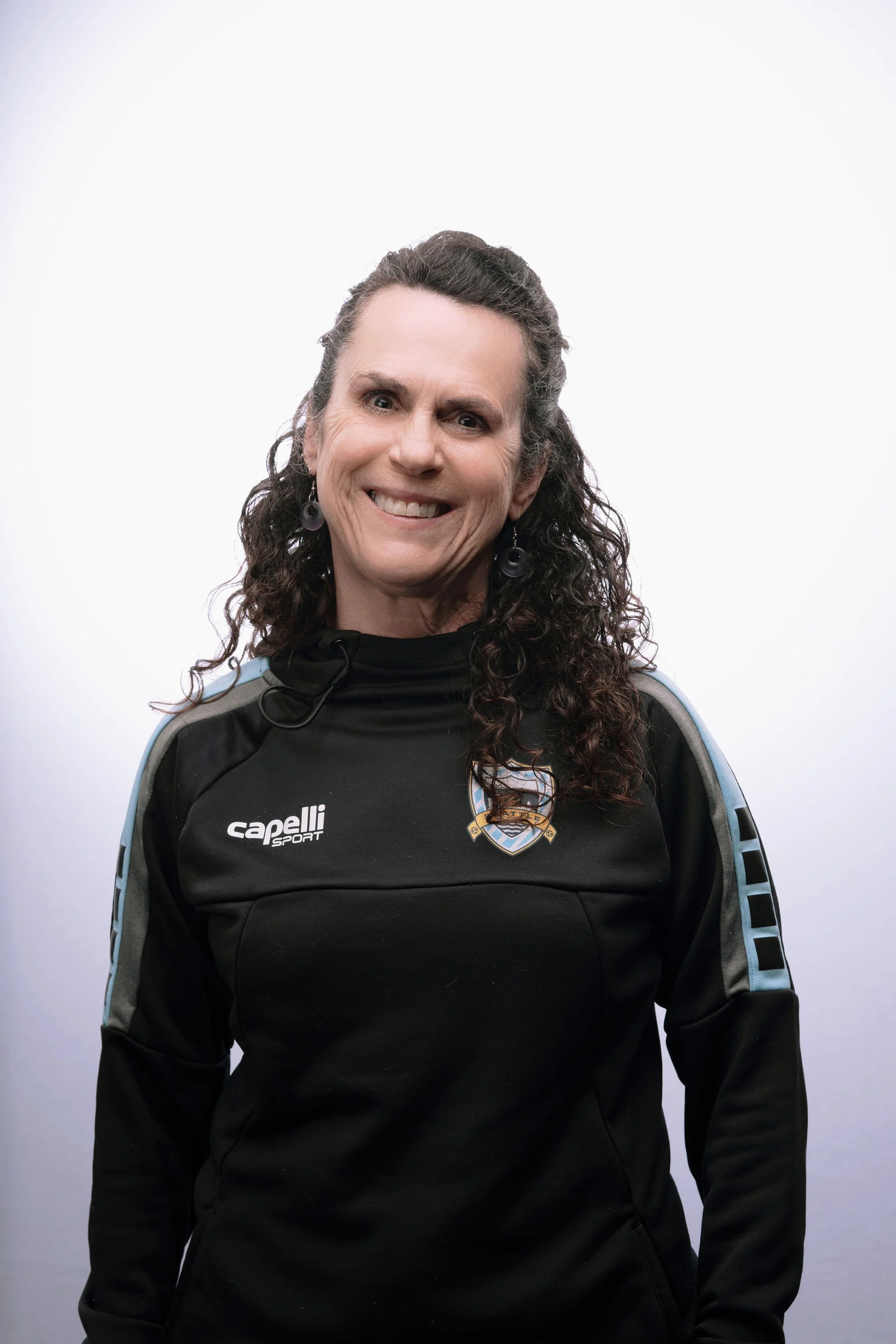 A woman with long curly hair smiling, wearing a black sports jacket with a logo and the brand name 'Capelli Sport' on it, against a plain background.