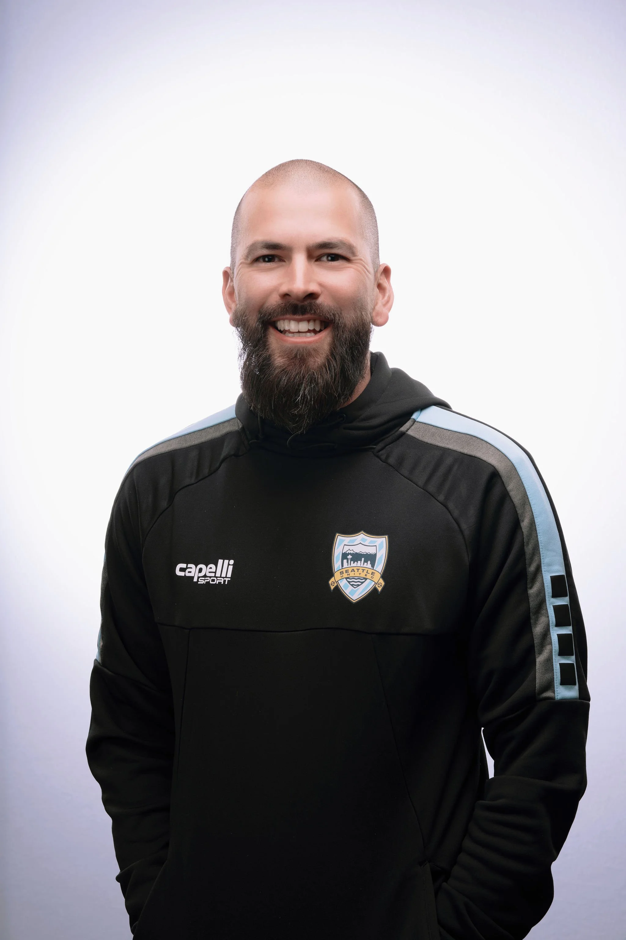A smiling man with a beard wearing a black Seattle Sounders hoodie, posing against a plain white background.