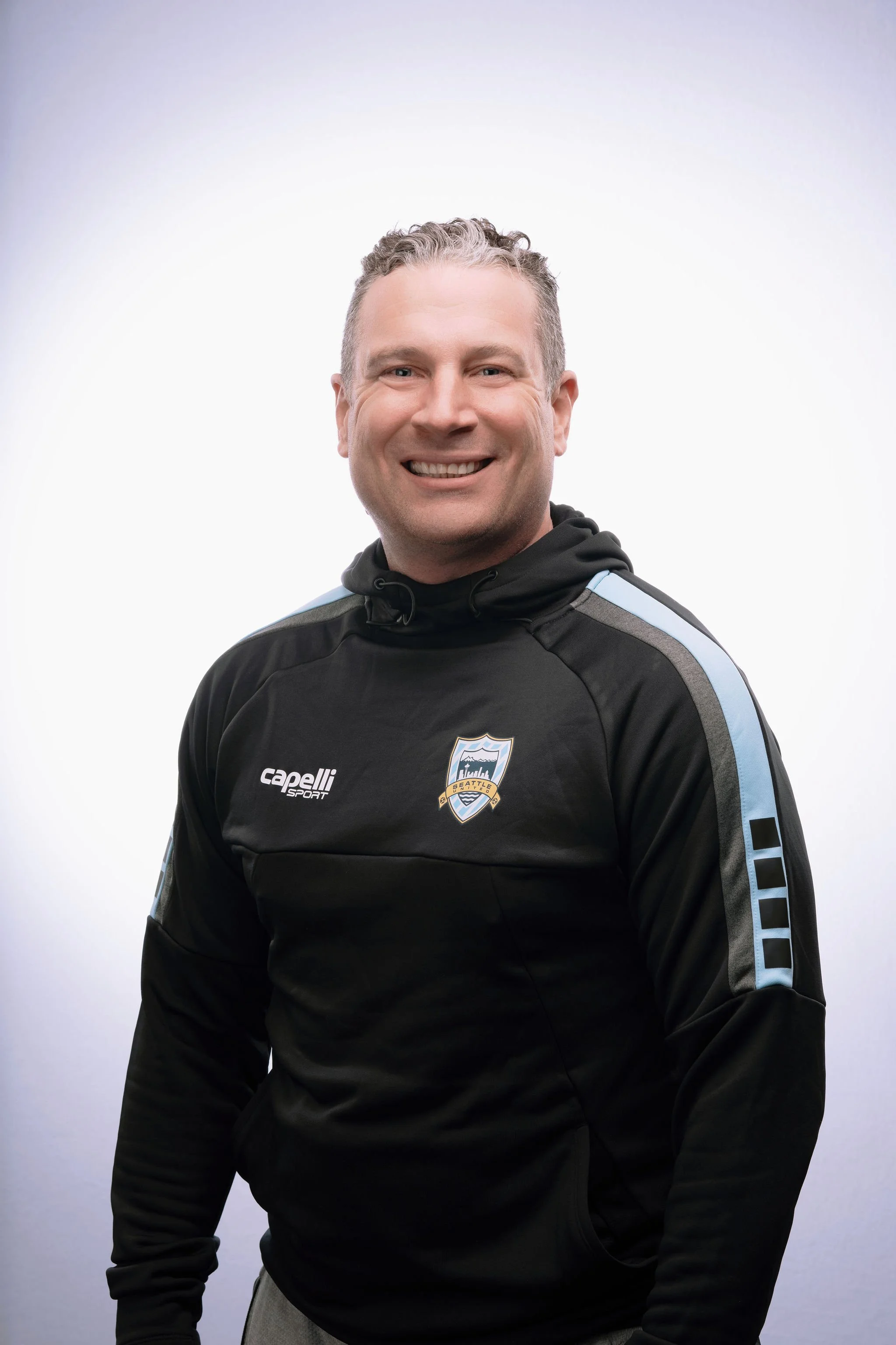 A man smiling in a black sports hoodie with the Seattle Sounders logo.