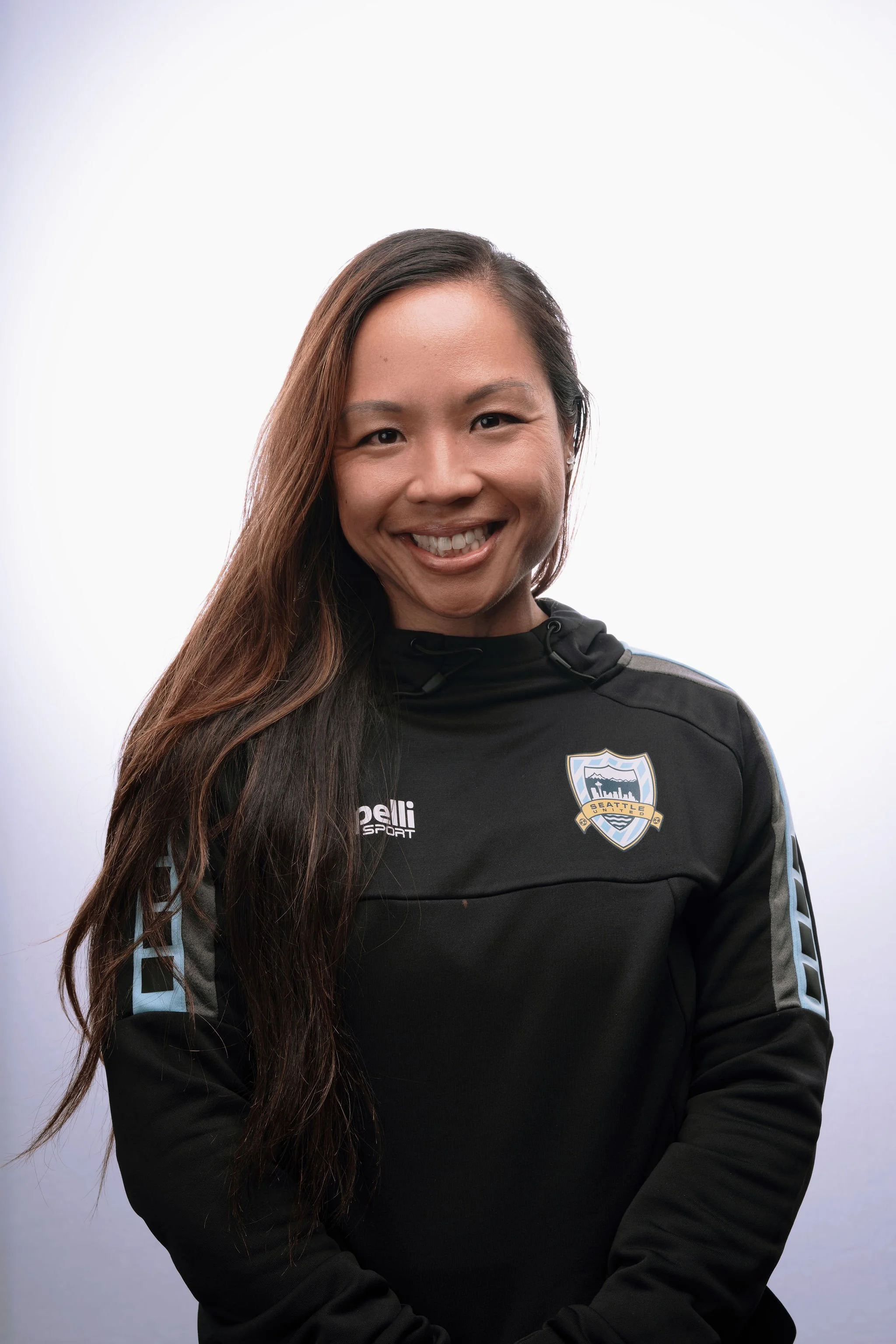 A woman with long dark hair smiling, wearing a black sports jacket with Seattle Sounders logo and sponsor text, standing against a plain background.