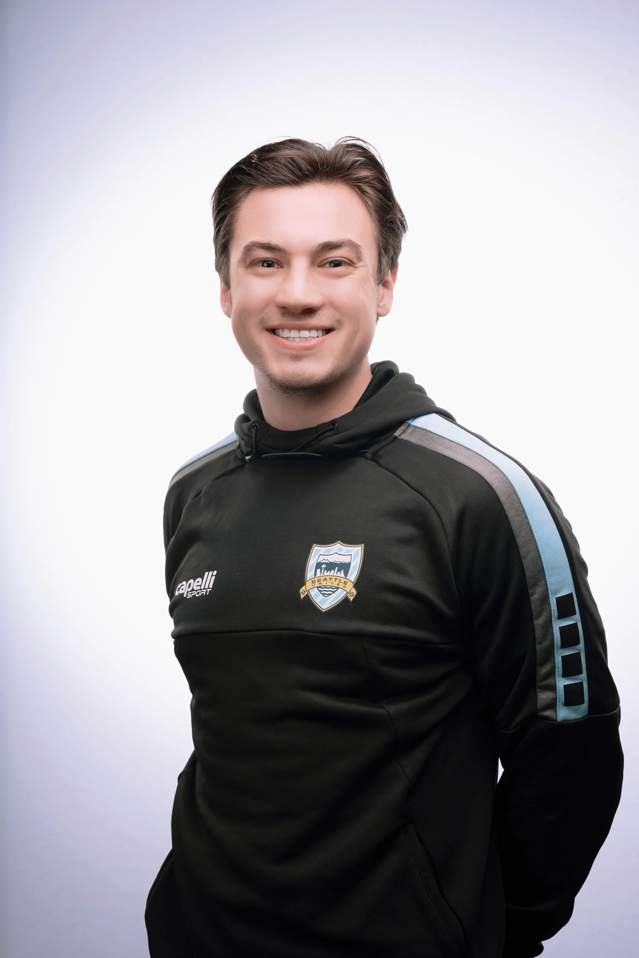 A young man smiling and wearing a black sports jacket with the Seattle Sounders logo on it, standing against a white background.