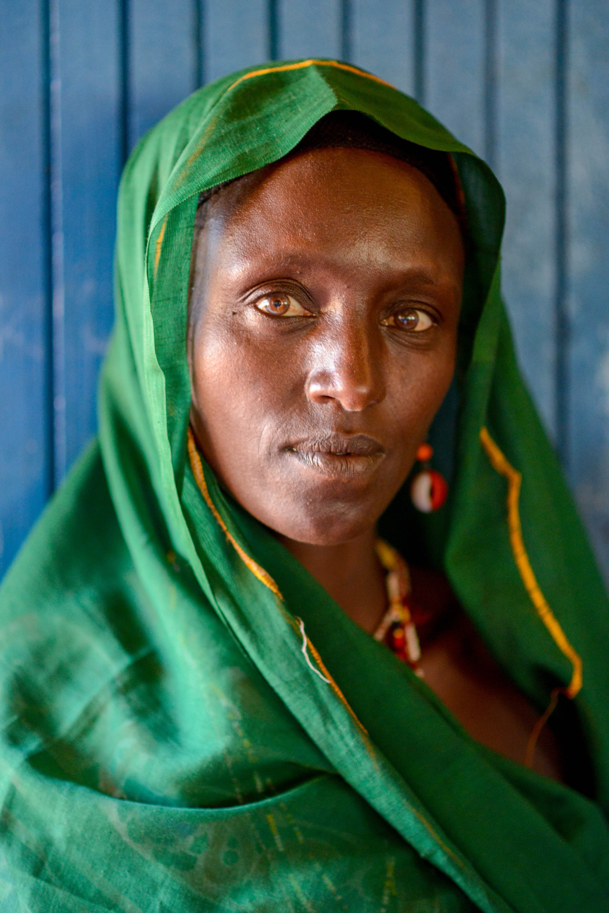 Close-up of a woman with dark skin wearing a green headscarf and traditional jewelry, standing against a blue wooden wall.