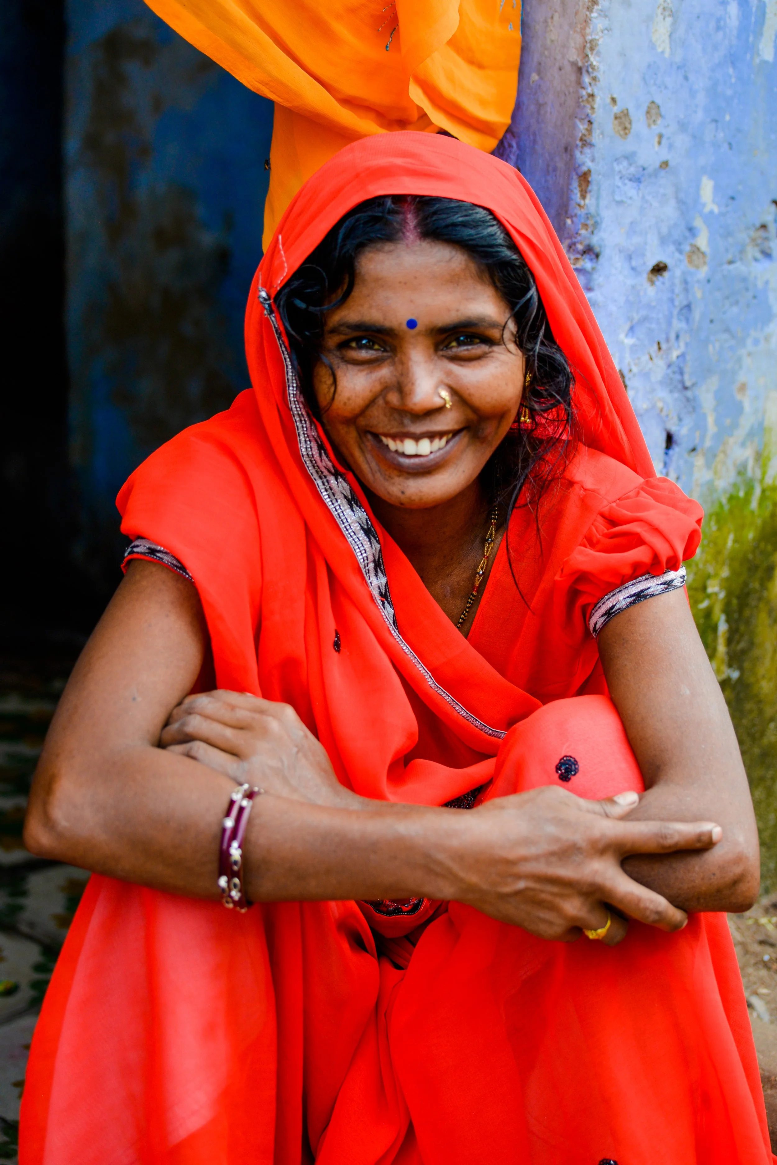 A smiling woman in India dressed in a bright red saree with a bindi on her forehead, sitting outdoors with a blue wall in the background.