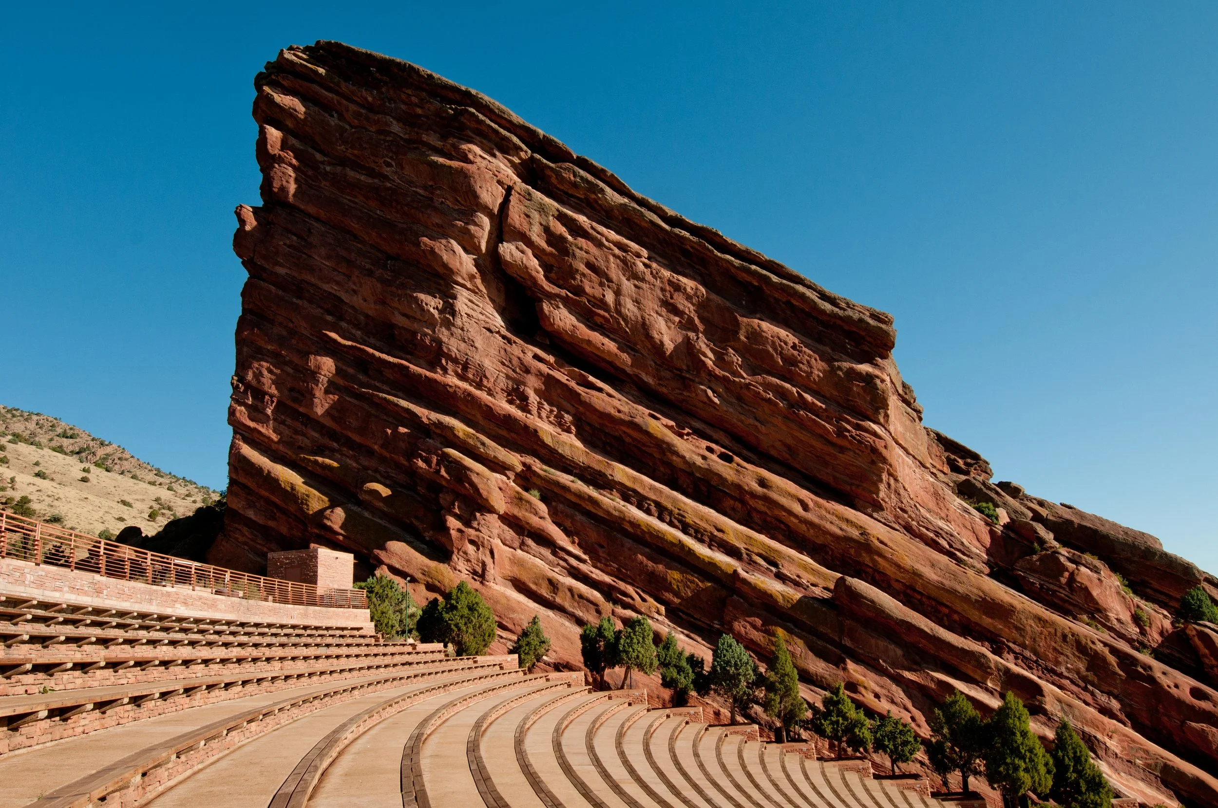 Red rock formation with tiered seating and small trees, under a clear blue sky.