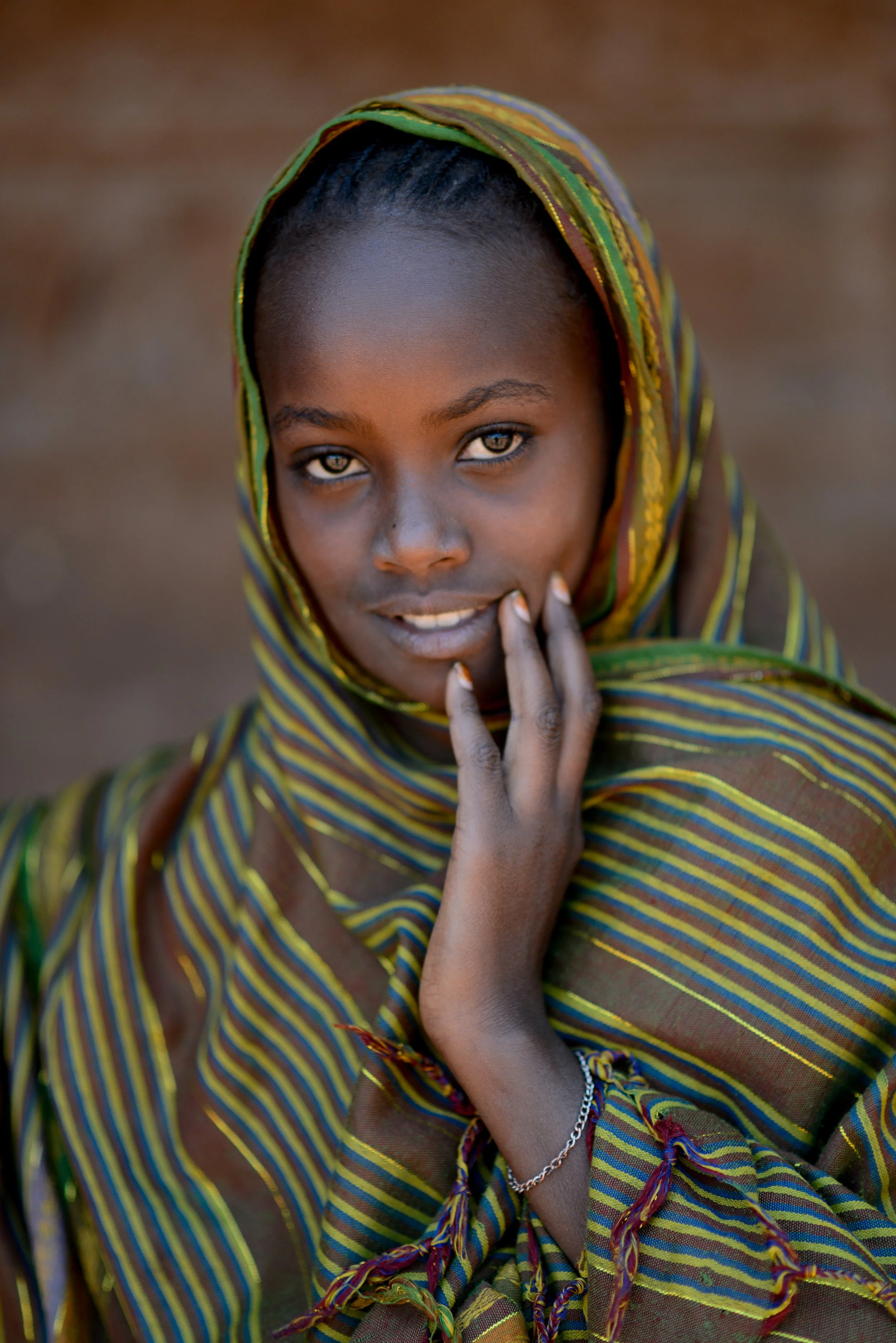 A young African woman wearing a colorful striped headscarf and shawl, touching her face with one hand, looking directly at the camera.