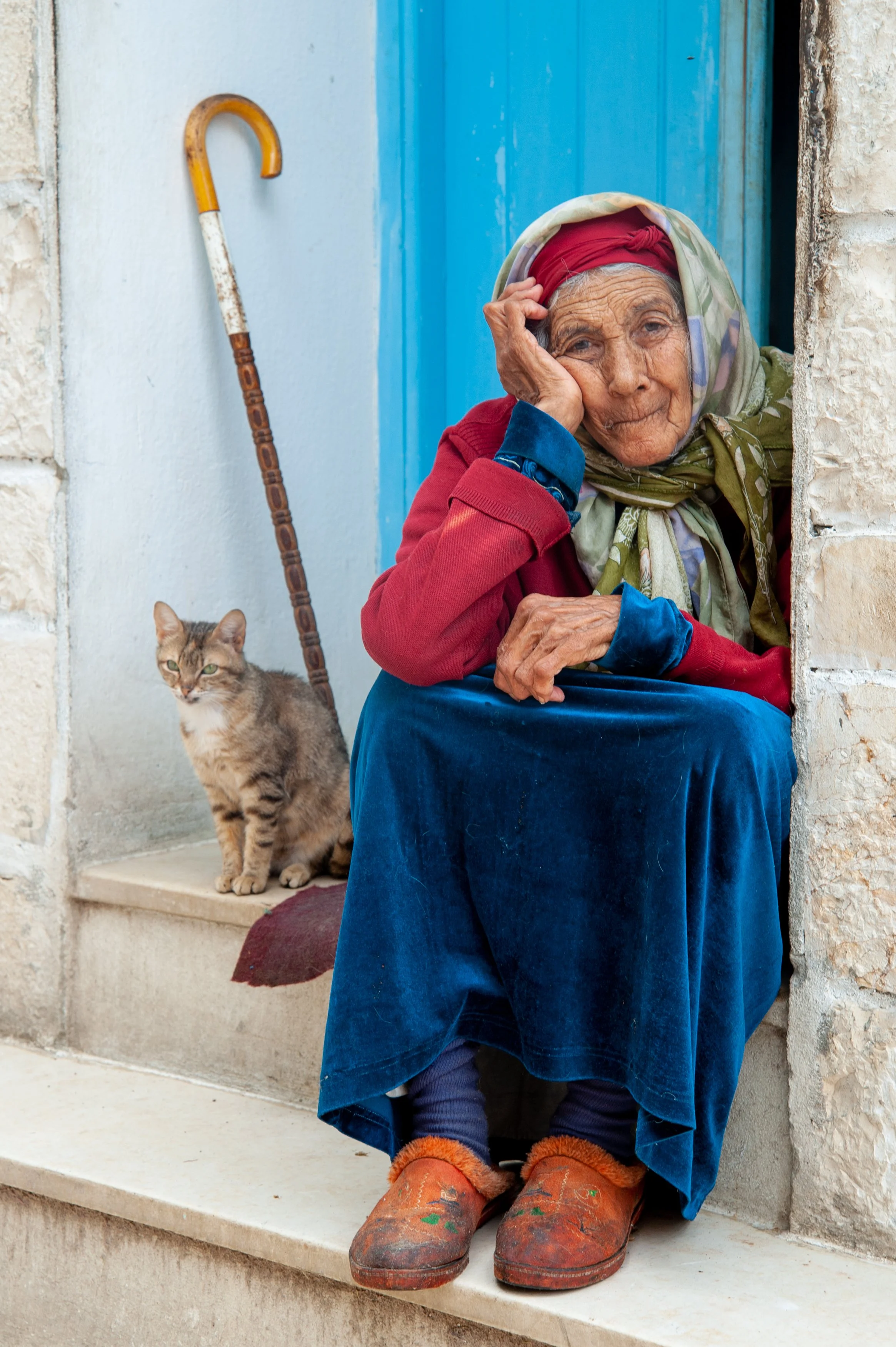 An elderly woman sitting at a doorway with a cat nearby, a cane behind her, dressed in colorful clothing and headscarf, looking contemplative.