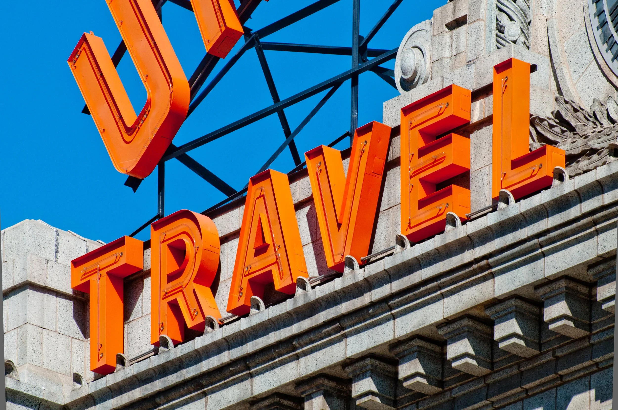 Close-up of a vintage orange neon sign spelling 'TRAVEL' mounted on a building with stone facade, under a clear blue sky.