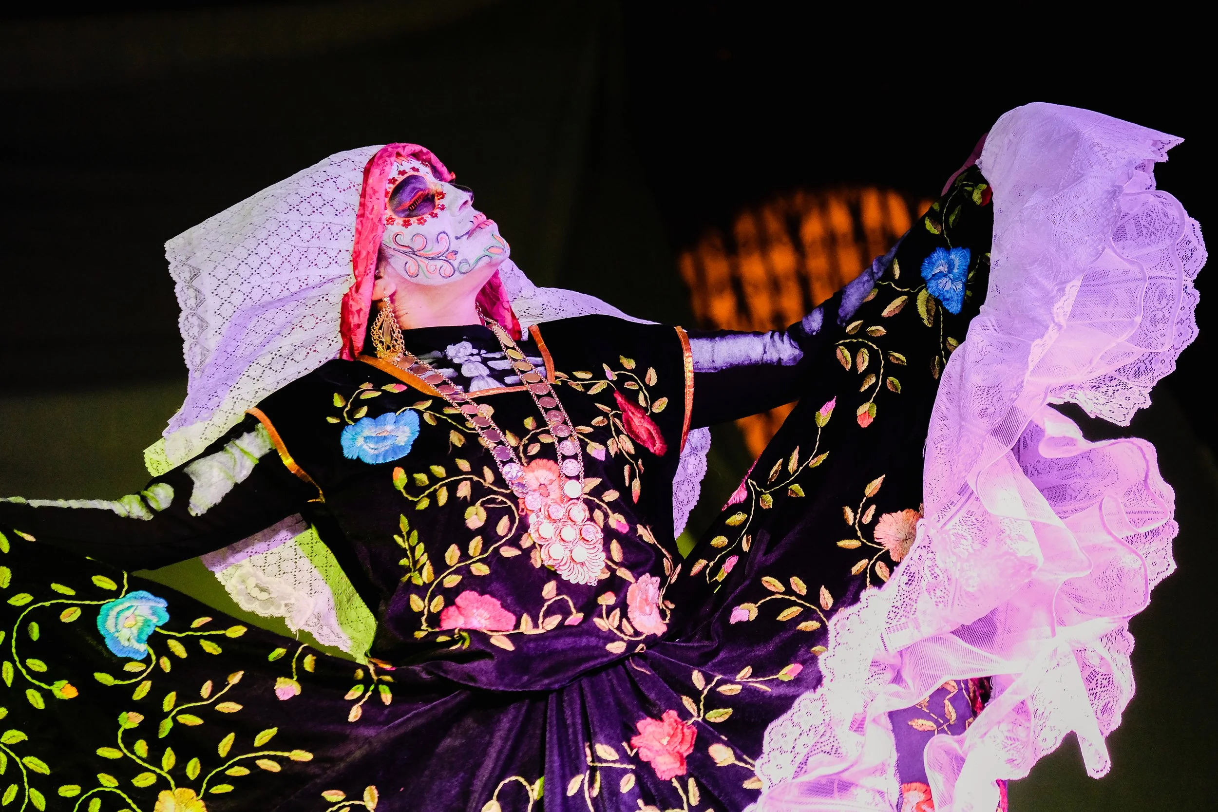 Traditional Mexican dancer wearing an embroidered black dress with floral patterns and lace details, with face paint, jewelry, and a veil, performing a dance on stage.