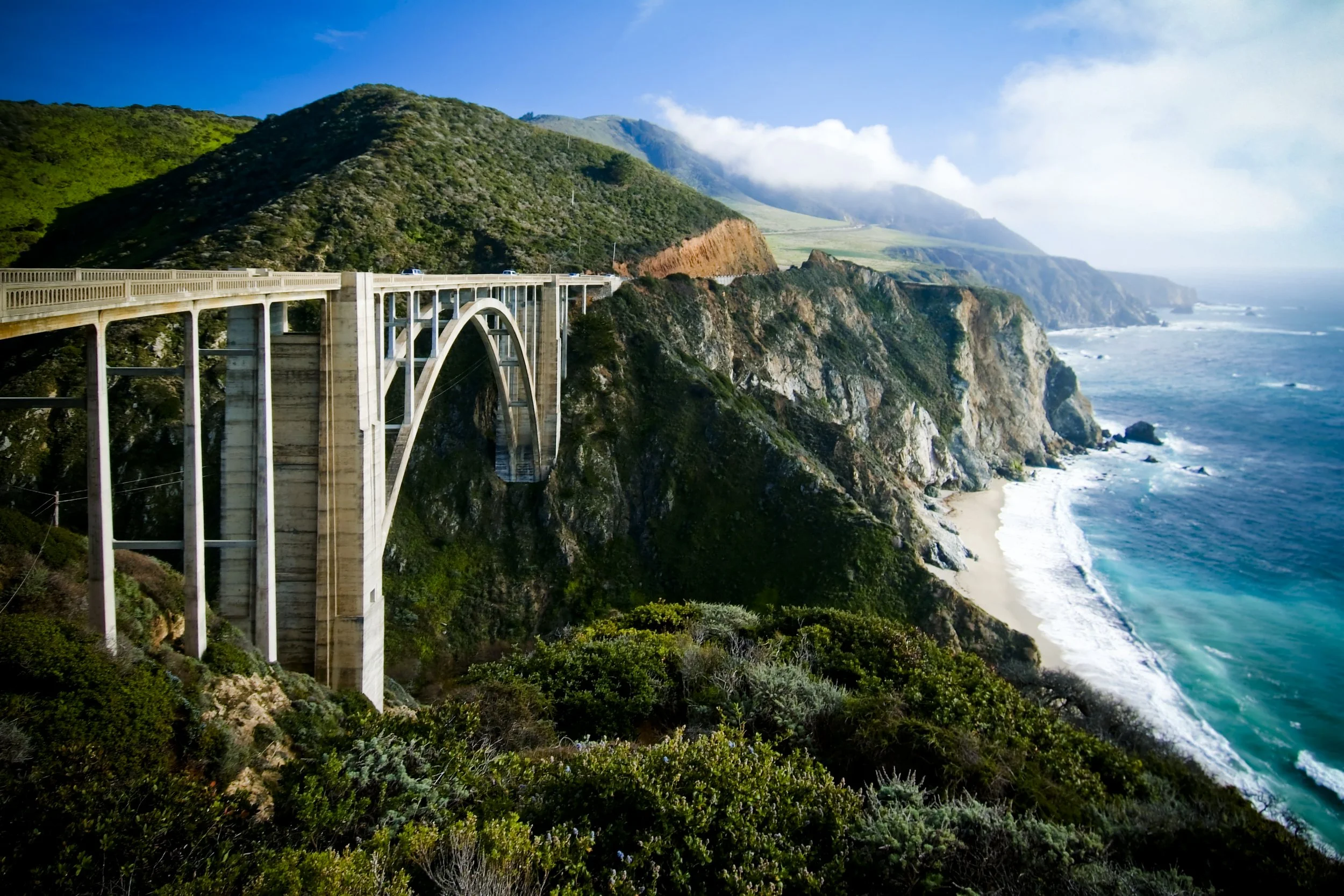 Photo of a coastal bridge over cliffs with ocean waves crashing on the beach underneath, green hills in the background, partly cloudy sky.
