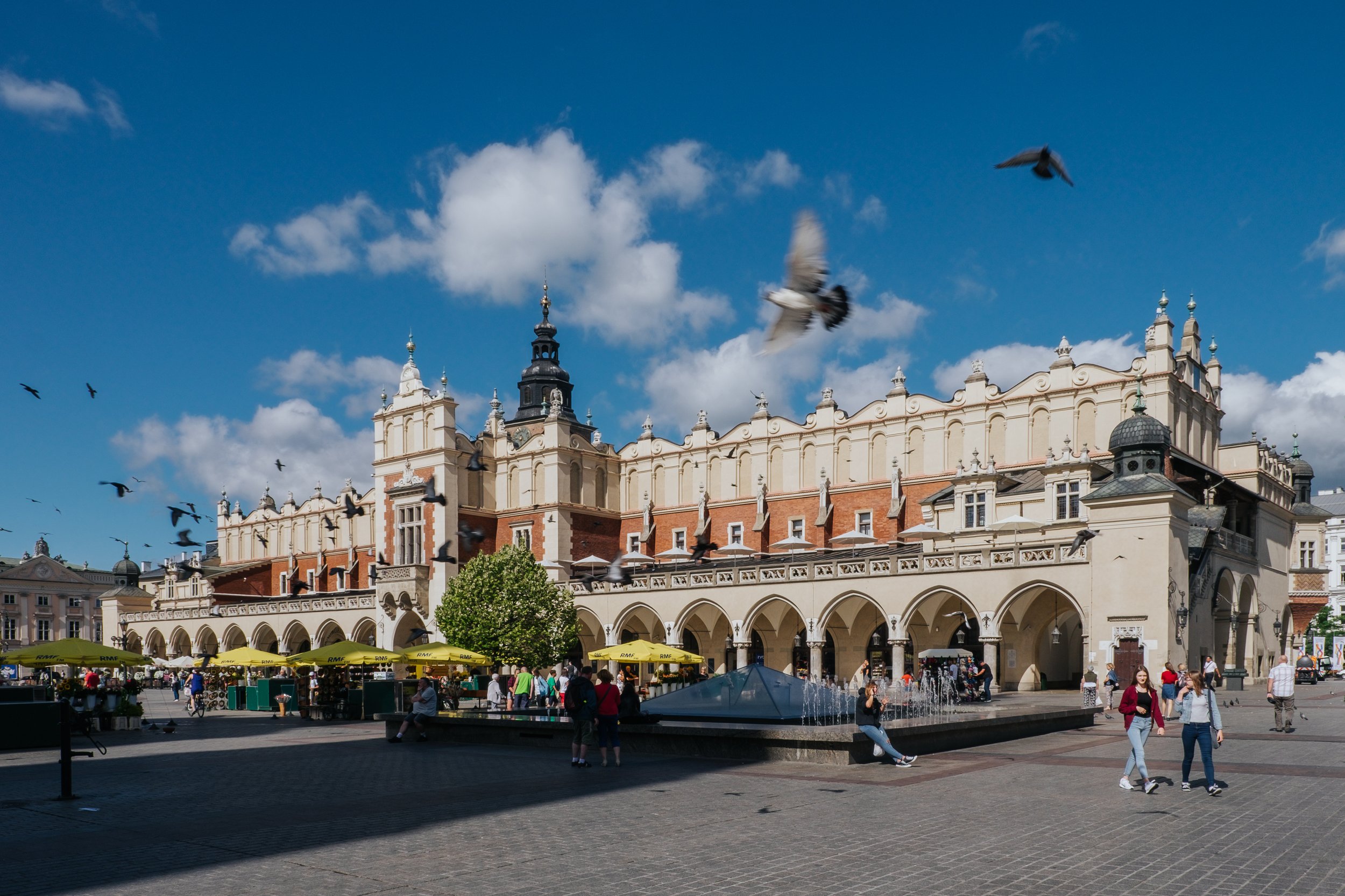 Historical building with arched entrance and tower in a busy city square, with pigeons flying overhead and people walking and sitting around, under a partly cloudy blue sky.