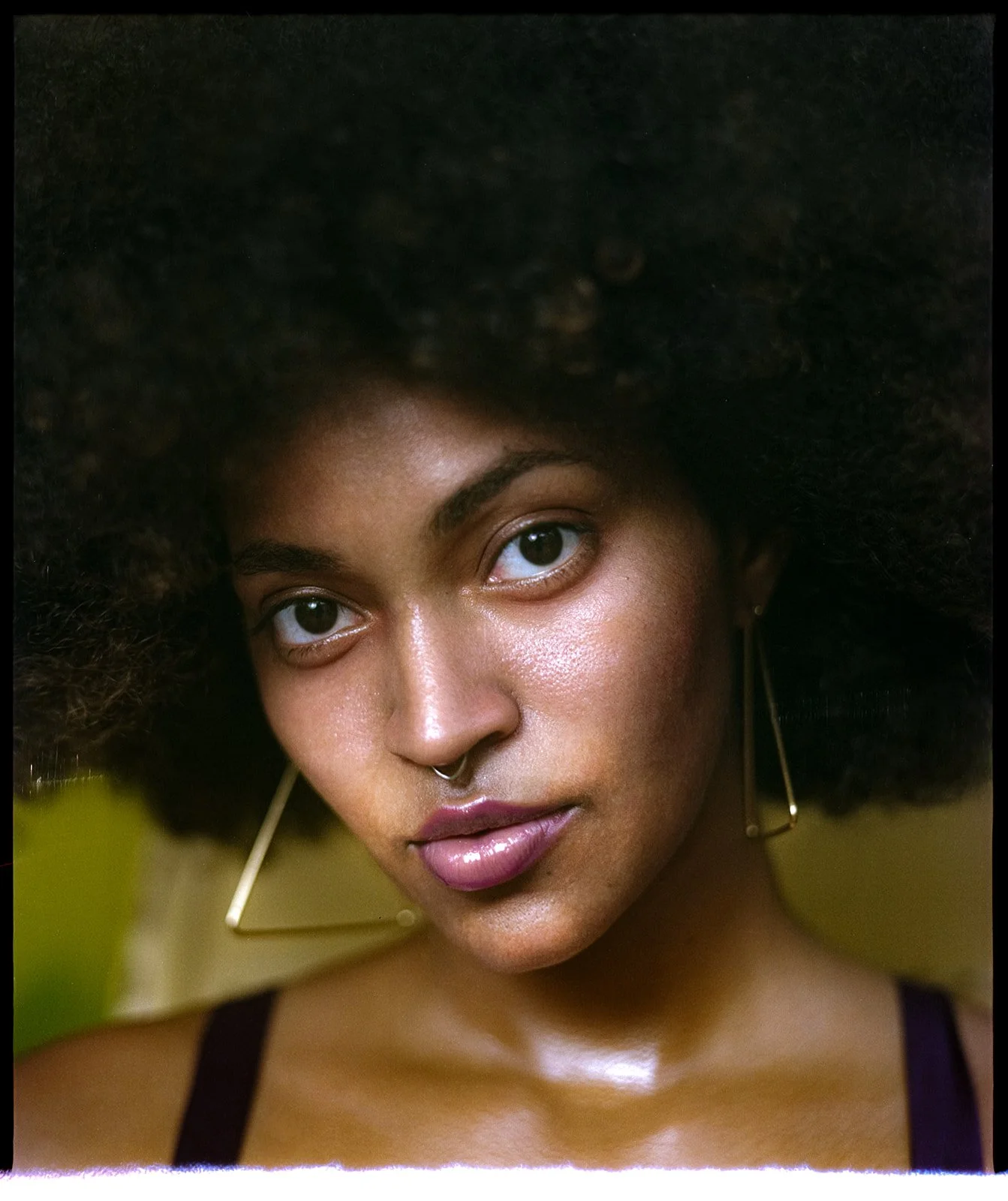 Close-up portrait of a woman with large curly afro hair, wearing a septum piercing, long triangle earrings, and dark clothing, with a blurred yellow-green background.