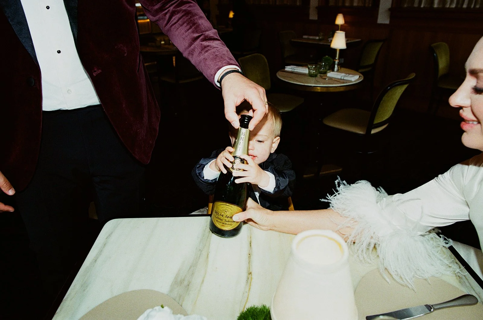 A young child in a dark jacket is sitting at a dining table, holding a champagne bottle with assistance from an adult. An adult woman with a white fur-lined sleeve is also reaching toward the bottle, smiling. The scene is in a dimly lit restaurant wi