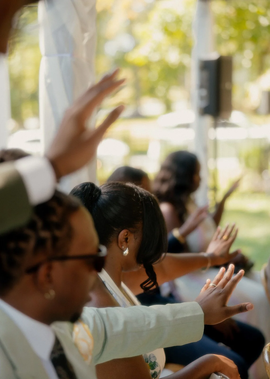 People seated with hands raised during a ceremony or event, outdoors with trees and sunlight in the background.