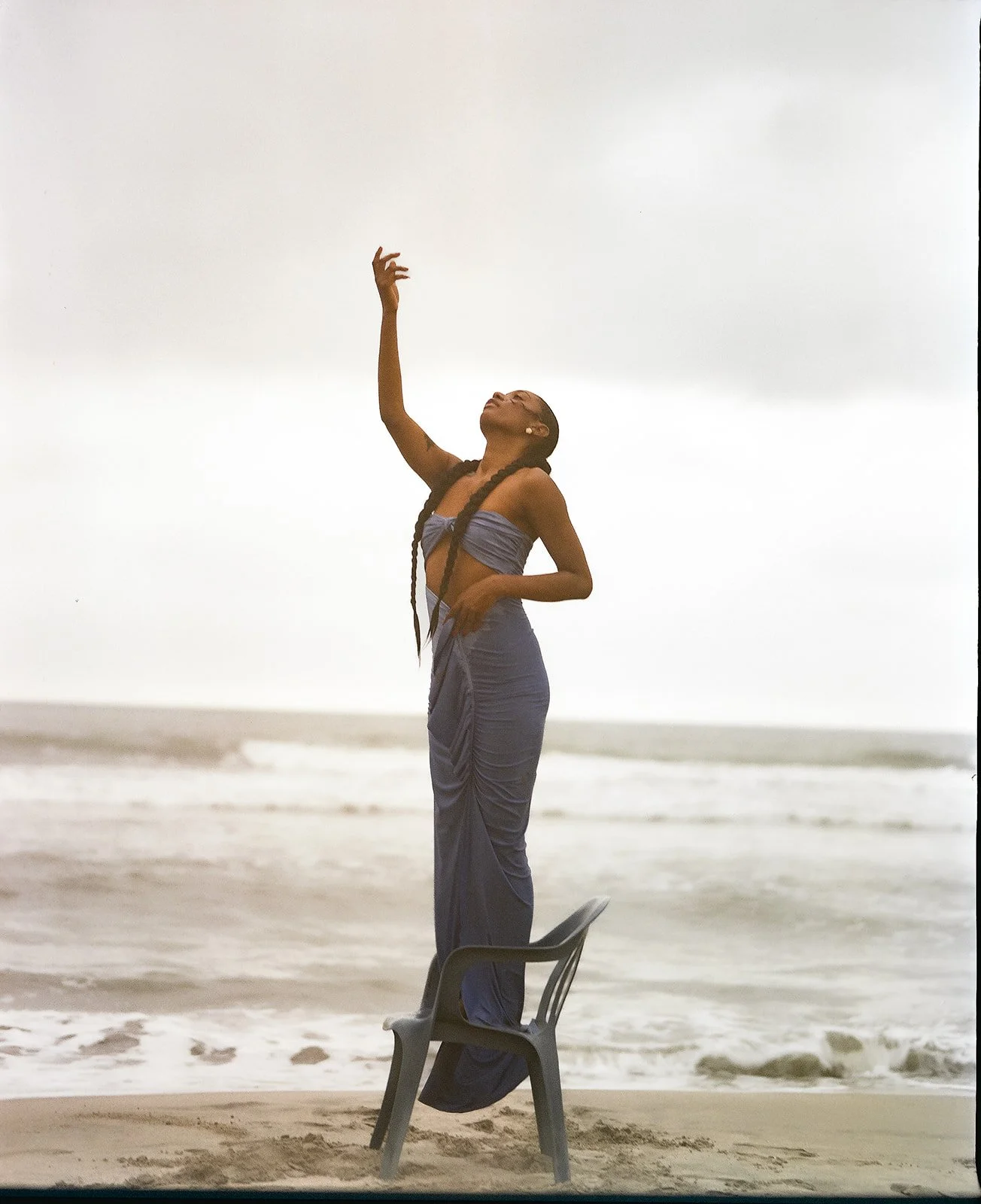 A woman in a blue outfit standing on a plastic chair on a beach, reaching upward with the ocean and cloudy sky in the background.