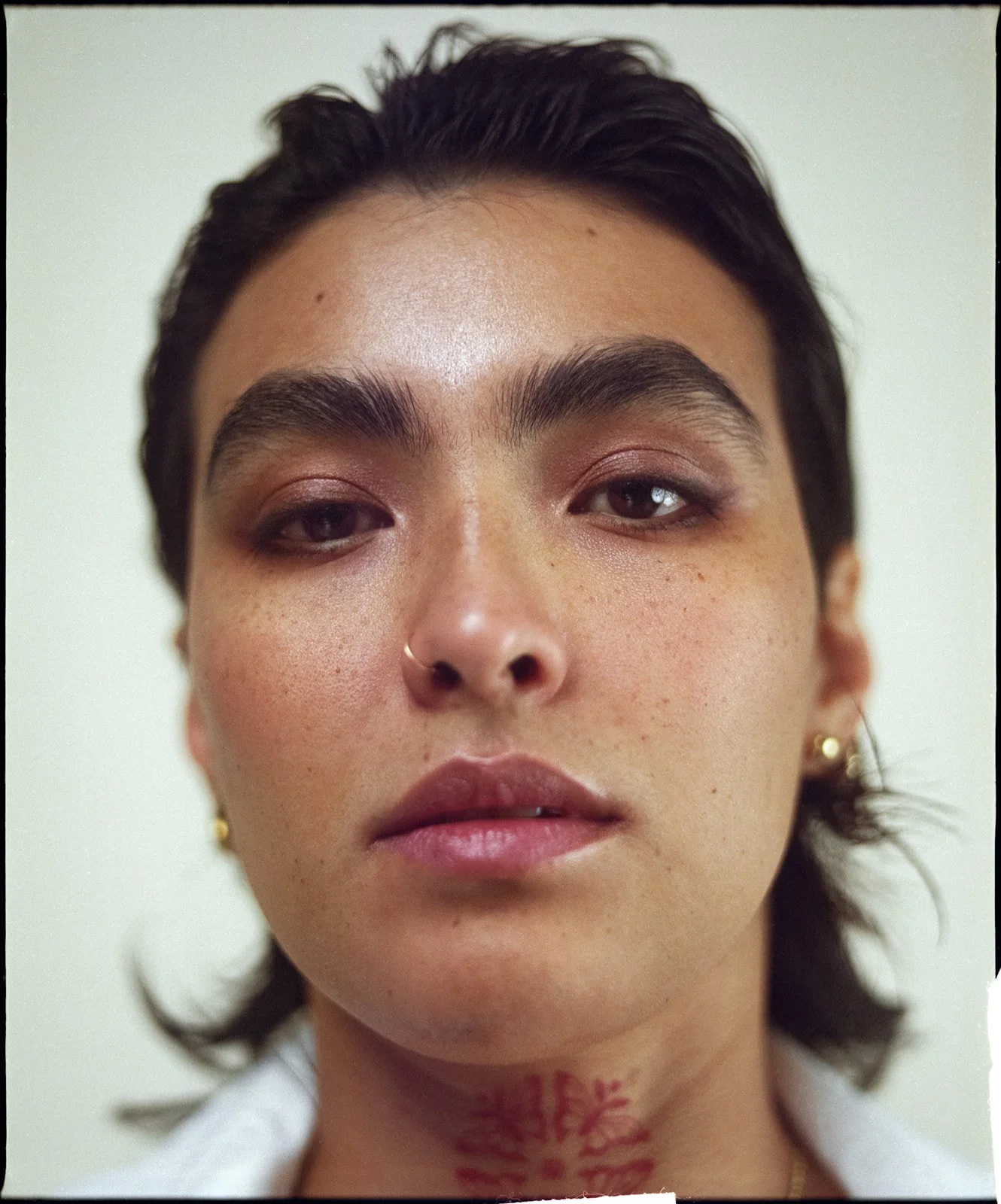 Close-up of a young person with dark hair, filled-in eyebrows, and minimal makeup, displaying a neck tattoo and wearing a white shirt.