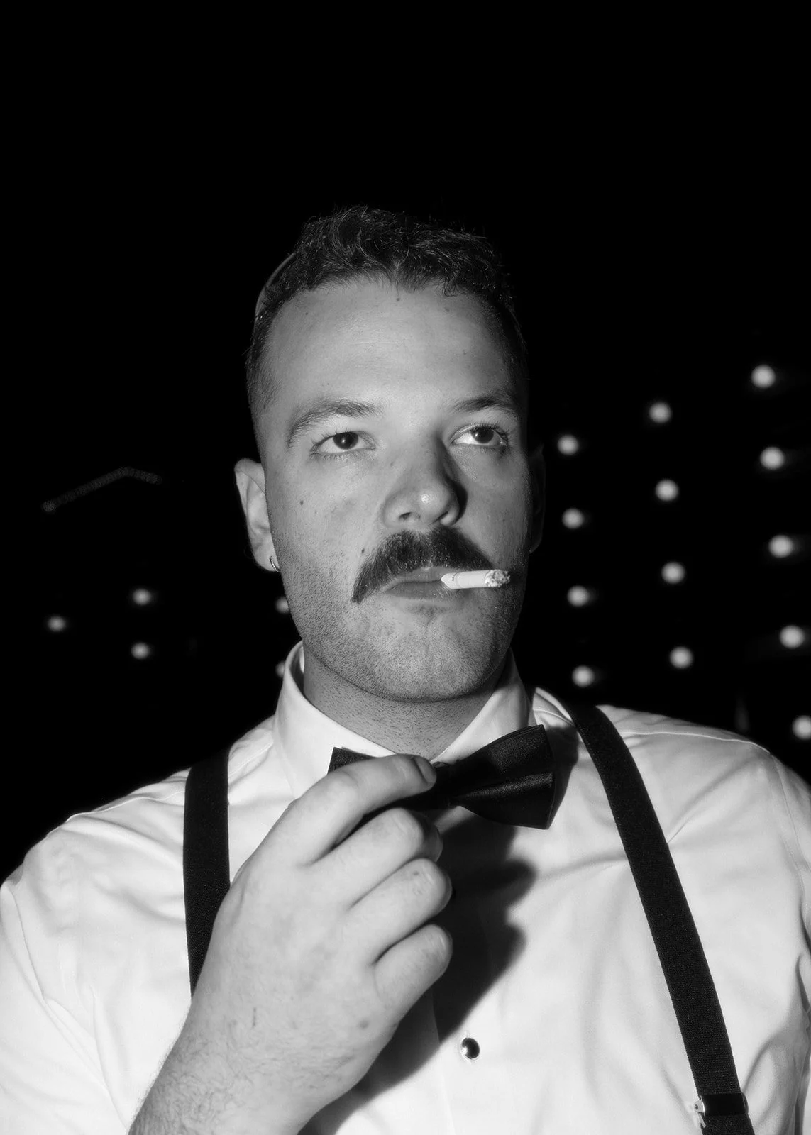 Black and white photo of a man with a mustache, dressed in a white shirt with suspenders and a bow tie, holding a cigarette in his mouth and adjusting his bow tie.