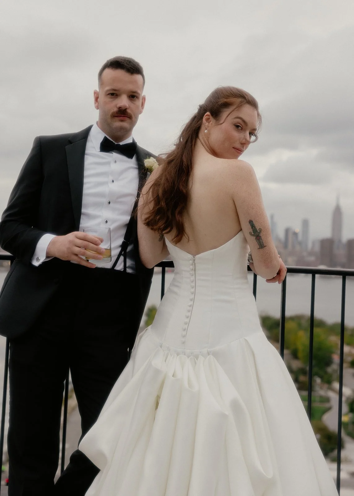 A newlywed couple on a balcony with a city skyline in the background. The groom is in a black tuxedo with a bow tie, holding a drink, and has a mustache. The bride is in a strapless white wedding gown with buttons down the back, revealing a tattoo of