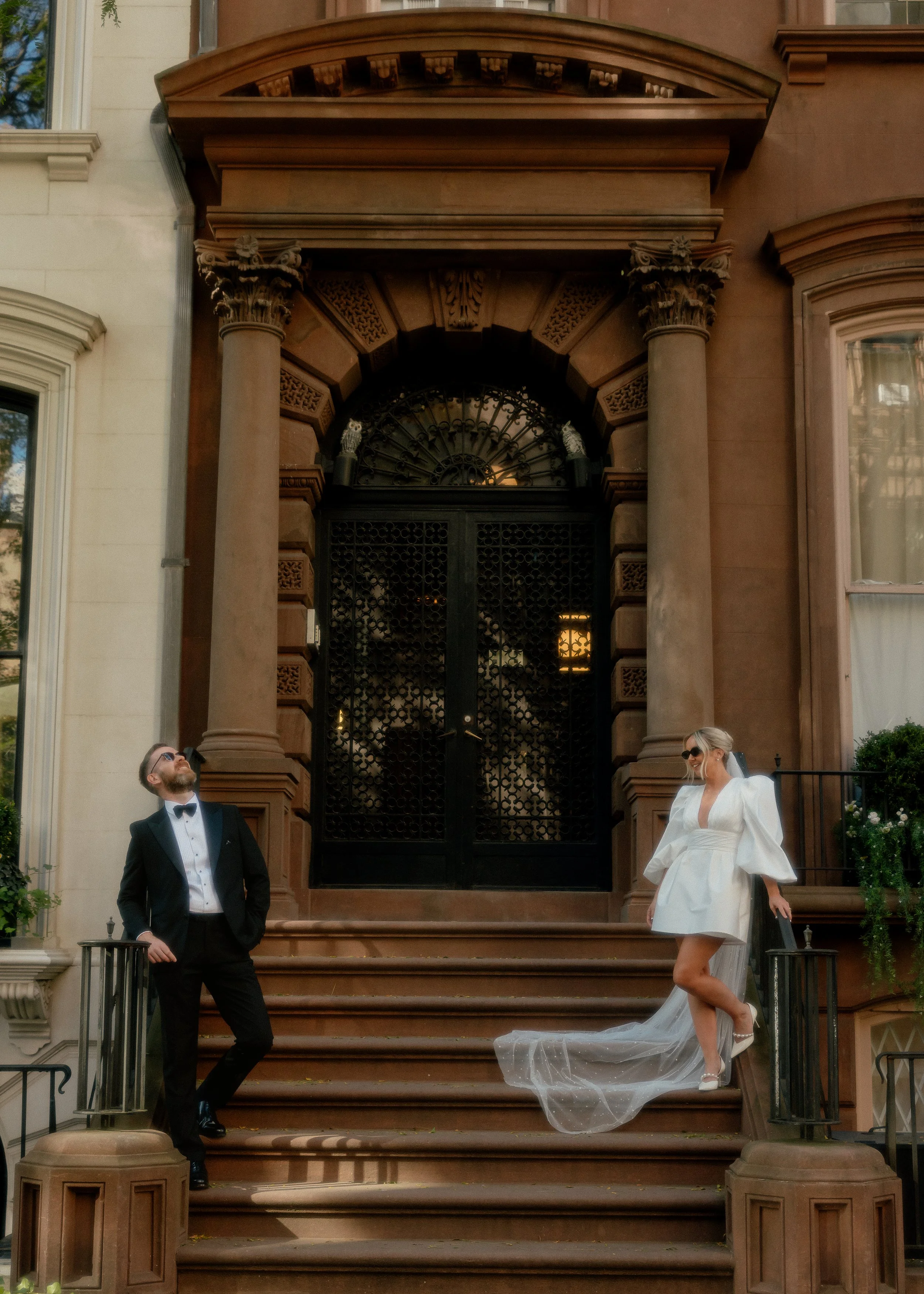 A newly married couple posing on the stairs outside a brownstone building. The groom is dressed in a black tuxedo with a bow tie, sunglasses, and has his hands in his pockets. The bride is wearing a white mini dress with puffed sleeves, high heels, a