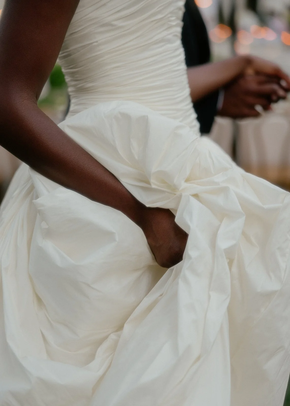 Close-up of a person wearing a white dress, adjusting the skirt, with another person in the background holding a cellphone.