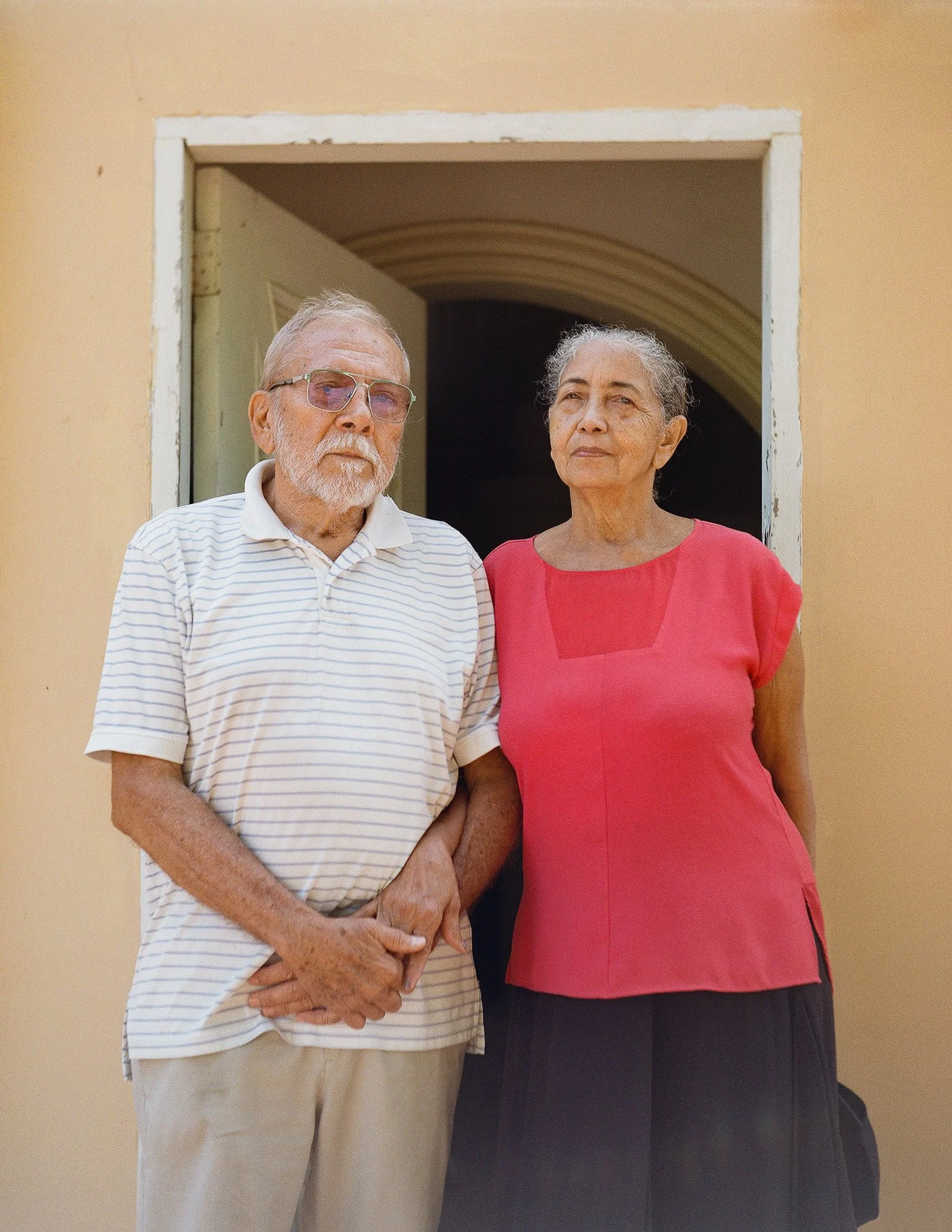 An elderly couple standing together in front of a doorway, the man with white hair and beard wearing glasses and a striped polo shirt, the woman with gray hair wearing a red blouse.