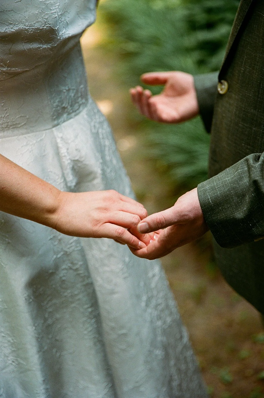 A couple holding hands during a wedding ceremony, with the bride wearing a silver textured dress and the groom in a dark suit.