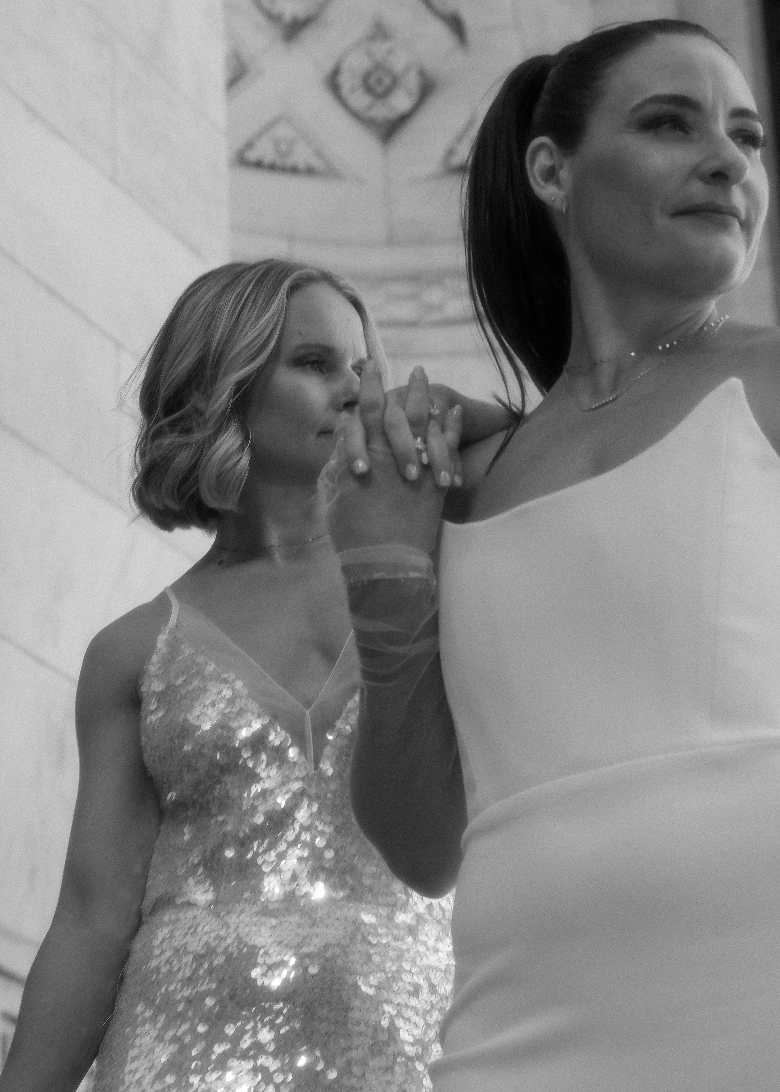 Two women, one with short curly hair in a sparkly dress and another with long dark hair in a white dress, holding hands in a prayer-like gesture, with the interior of a building in the background.