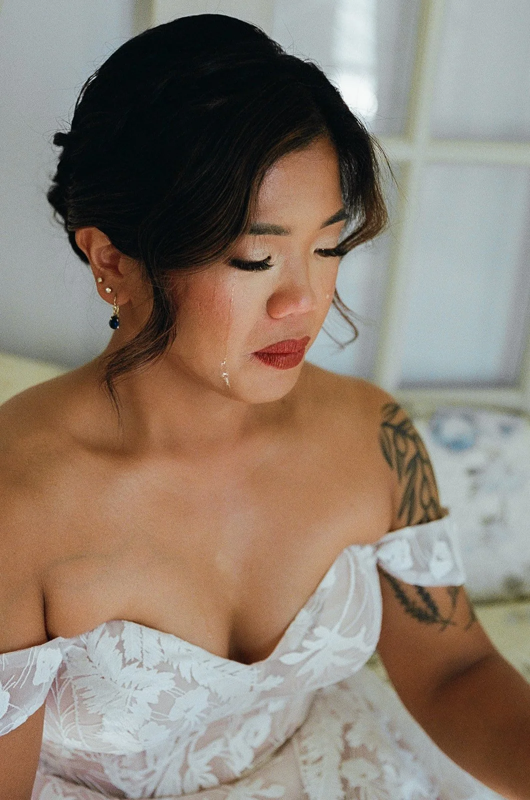 A woman with dark hair, earrings, and a tattoo on her shoulder, wearing an off-shoulder white dress, with tears on her face.