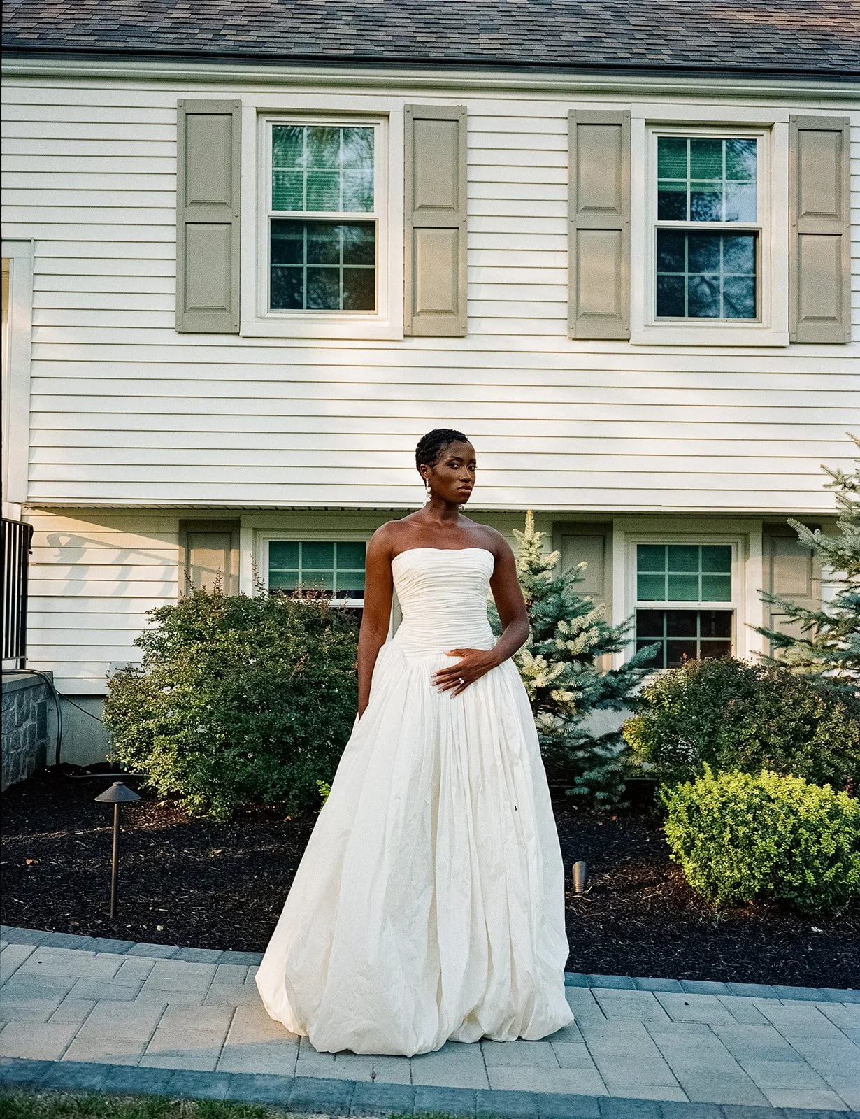 A woman in a strapless white wedding dress standing outside in front of a house with white siding and bushes.