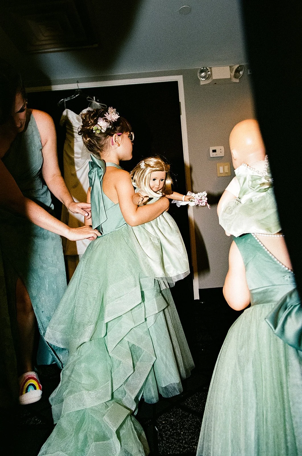 Children in green dresses getting ready, including one girl holding a doll, in a room with dark walls.